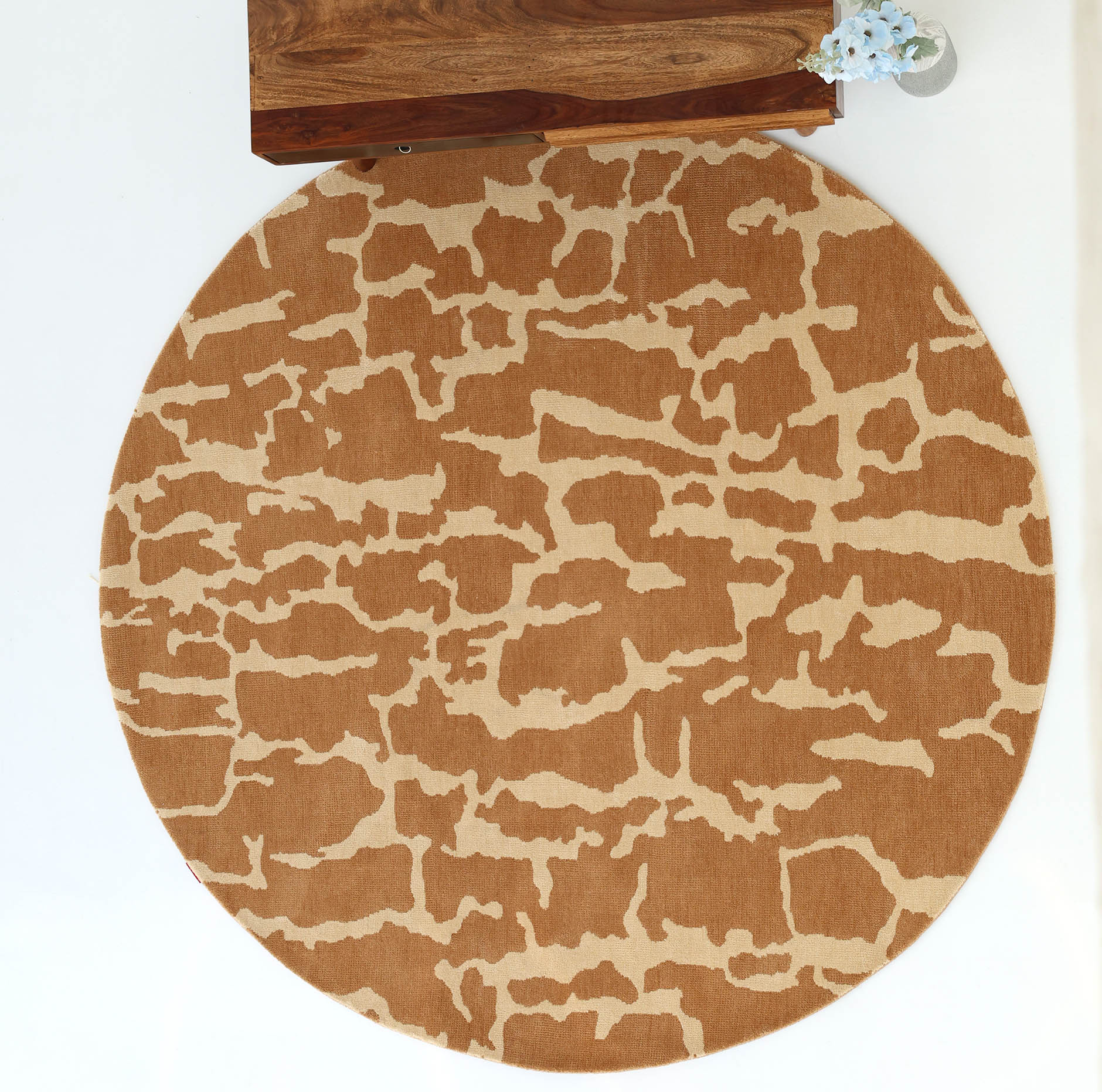 The RAMSHA HOME Natural Drift Round Area Rug CMN 116, featuring a brown and beige abstract pattern, rests on a white floor. In the top corner, part of a wooden table with a small vase of white flowers is visible.