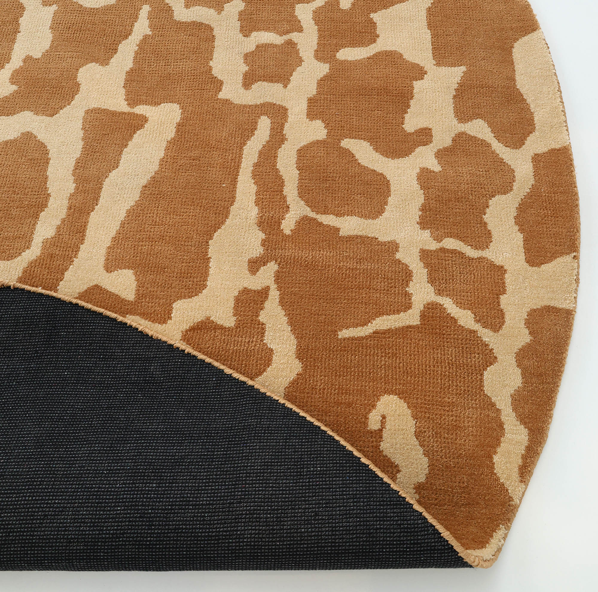 The RAMSHA HOME Natural Drift Round Area Rug CMN 116, featuring a modern brown and beige giraffe-like pattern, is partially folded to show its black underside against a white background.