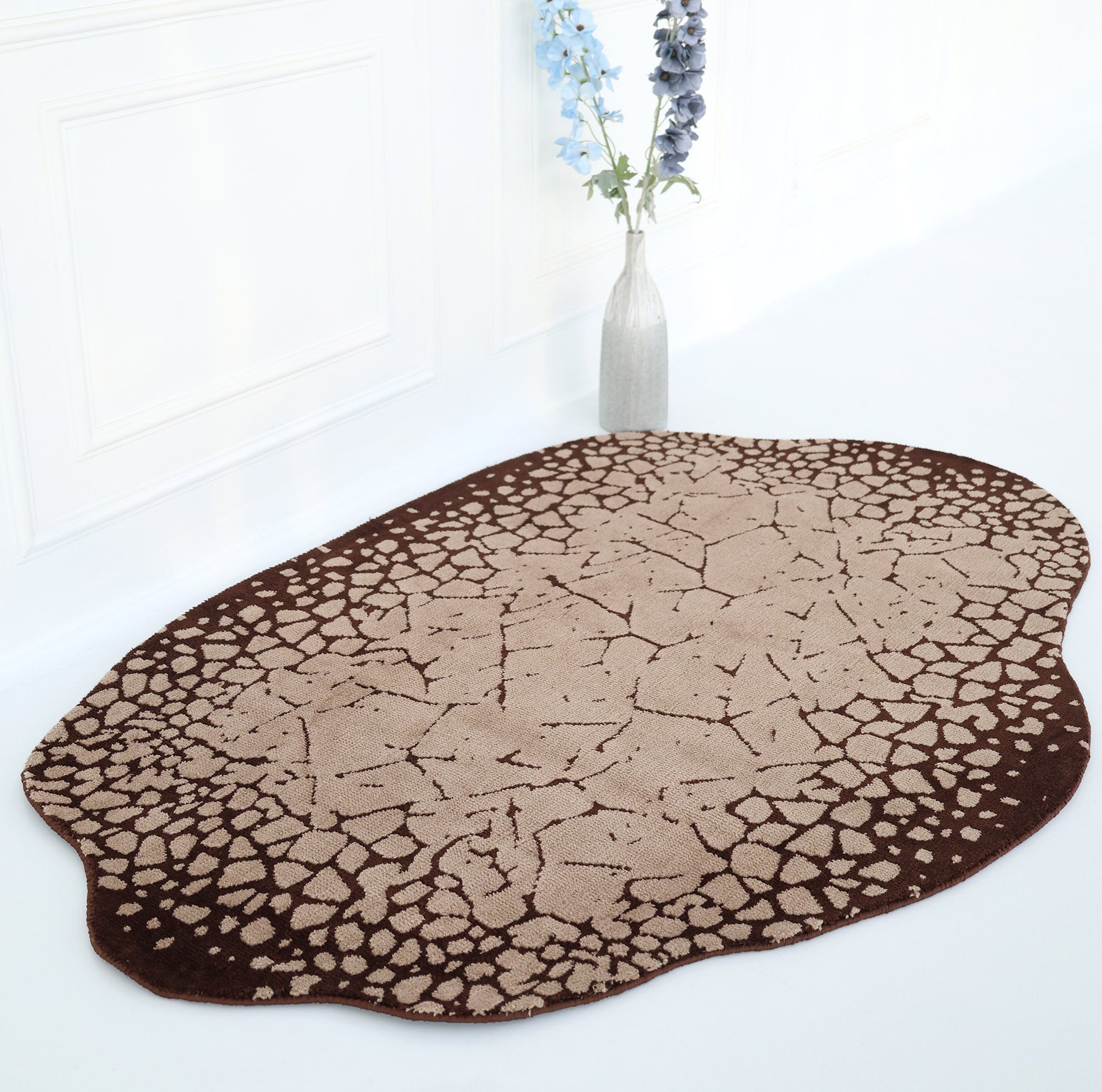 The Terrain Echo Abstract Rug Shaped Rug CMN 118 by RAMSHA HOME, featuring a brown and beige cracked earth pattern, is displayed on a white floor near a white wall with a tall vase of blue and purple flowers in the background.