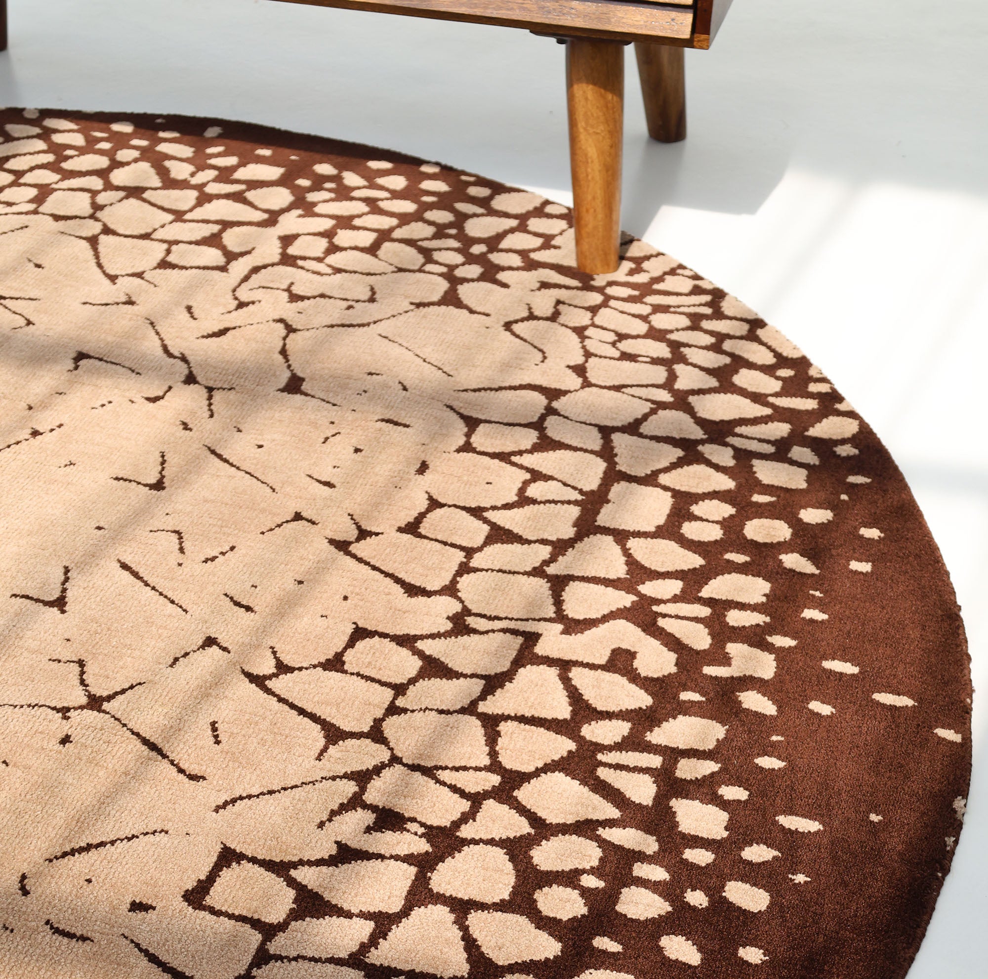 The RAMSHA HOME Terrain Echo Abstract Rug Shaped Rug CMN 118, featuring a beige and brown cracked-earth geometric pattern, sits under wooden furniture on a light floor as sunlight creates soft shadows in the modern space.