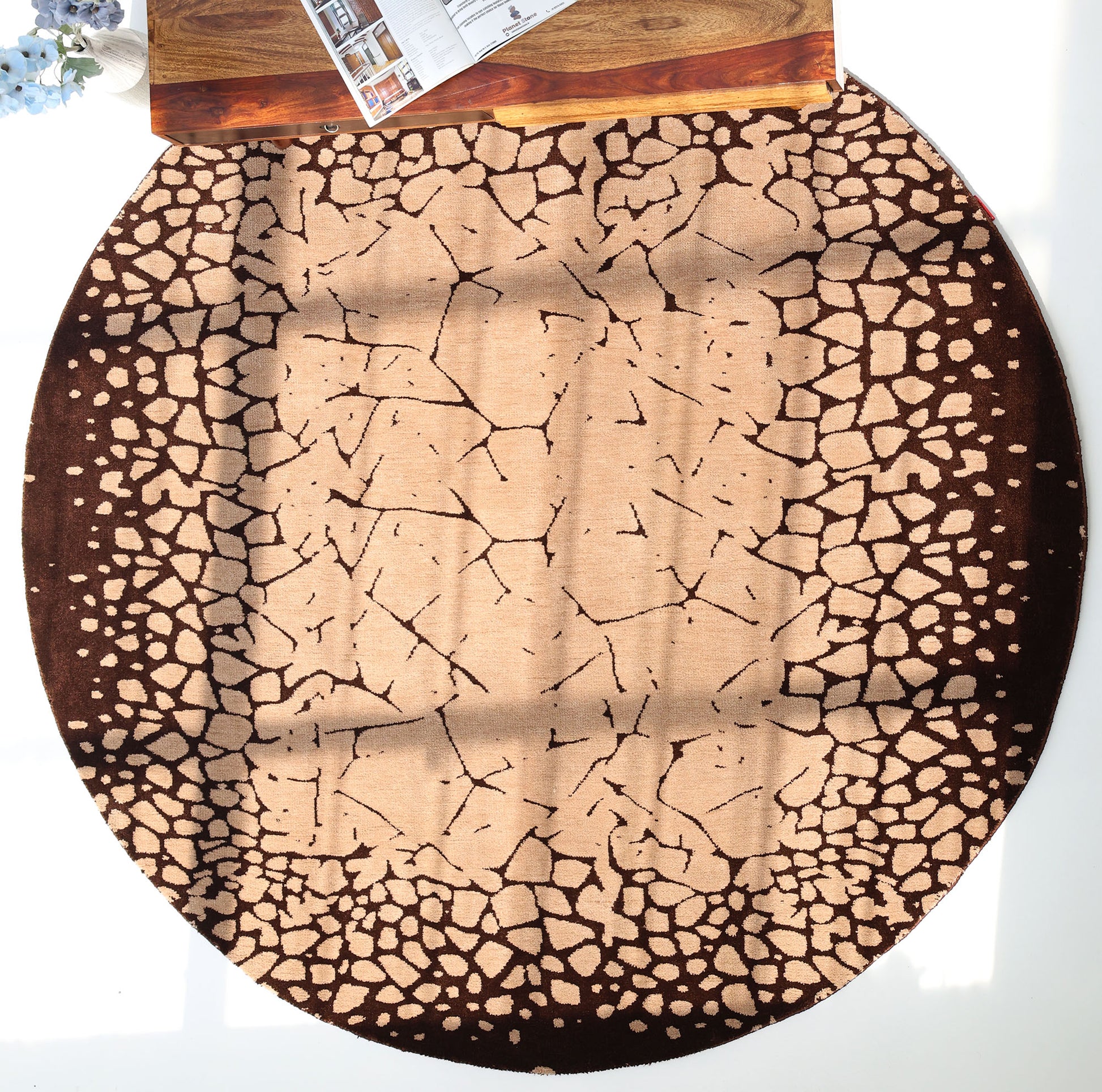 The RAMSHA HOME Terrain Echo Abstract Rug Shaped Rug CMN 118, featuring a cracked earth pattern in beige and brown, rests on a white floor by a wooden table holding a newspaper and blue flowers as sunlight casts shadows across its modern design.