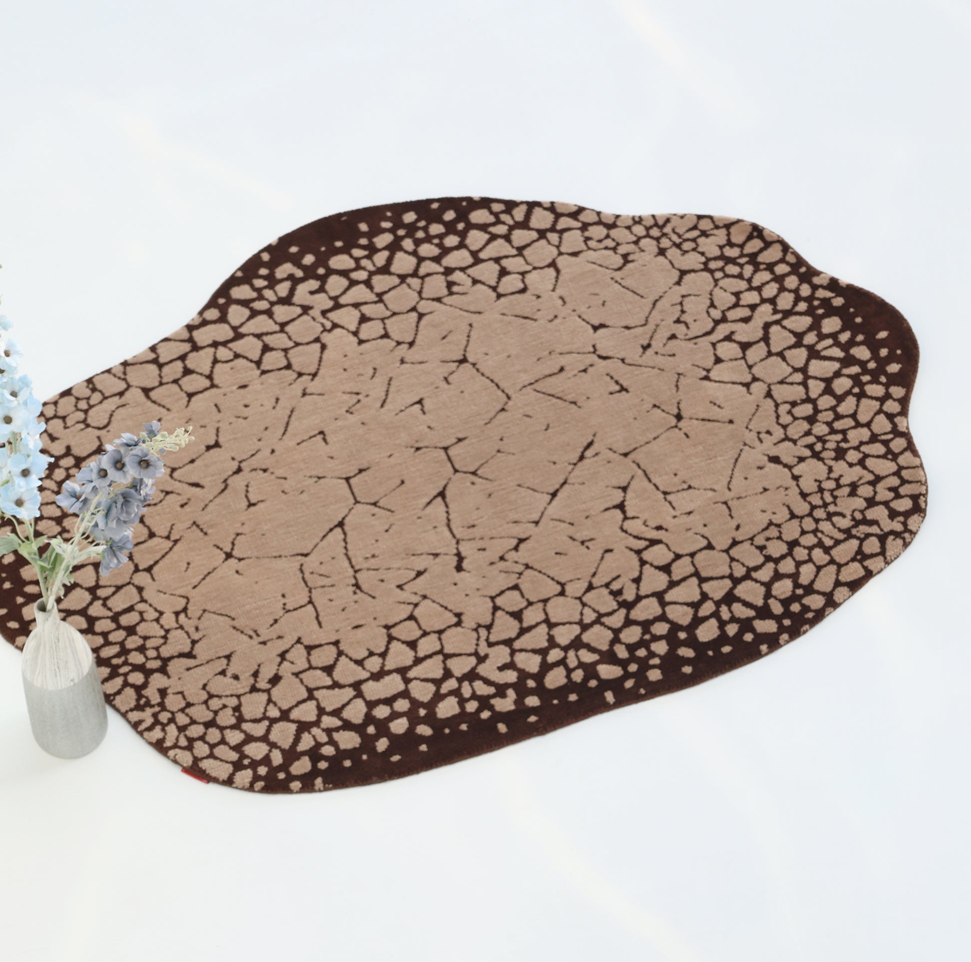 The RAMSHA HOME Terrain Echo Abstract Rug CMN 118, featuring a cracked earth pattern in beige and brown, lies on a white floor next to a white vase with lavender and blue flowers.