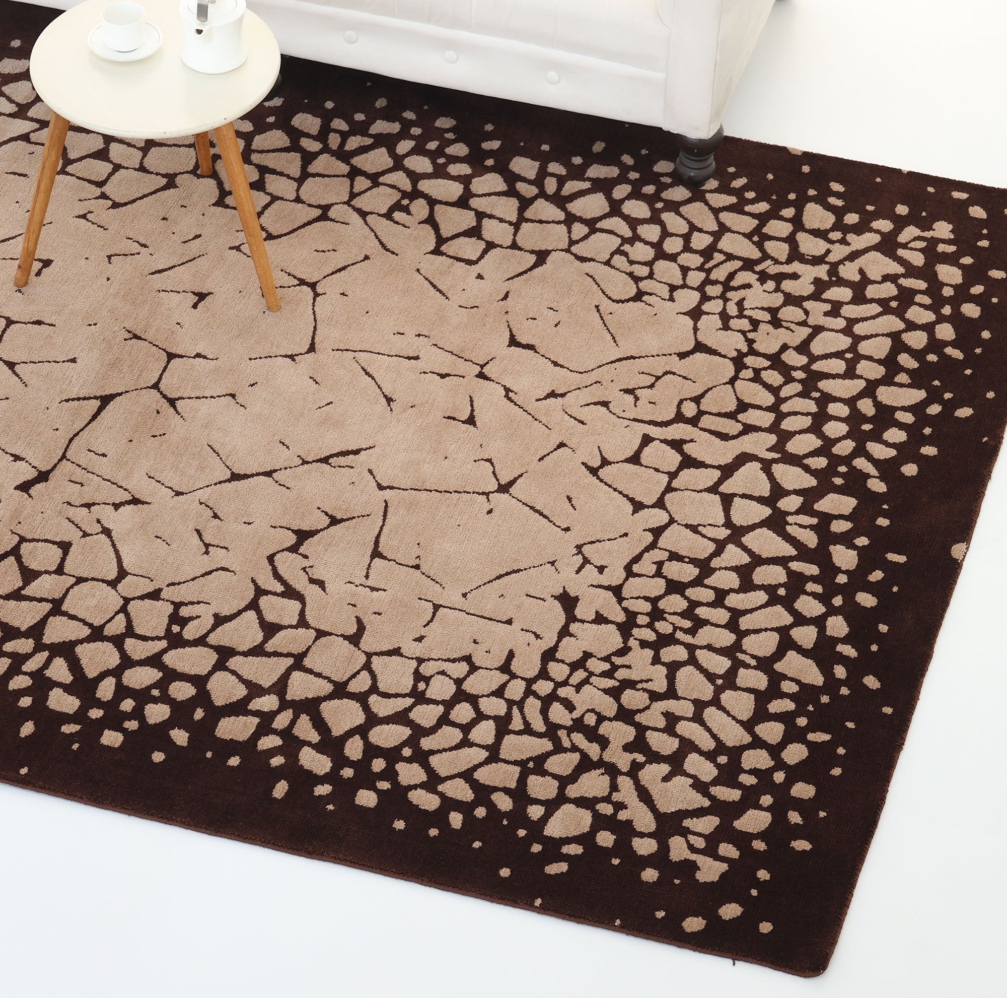 The Terrain Echo Abstract Rug Shaped Rug CMN 118 by RAMSHA HOME features a square design with scattered stone patterns, a beige center, and dark brown border. Part of a white sofa and round table with cups appear in the upper left corner.
