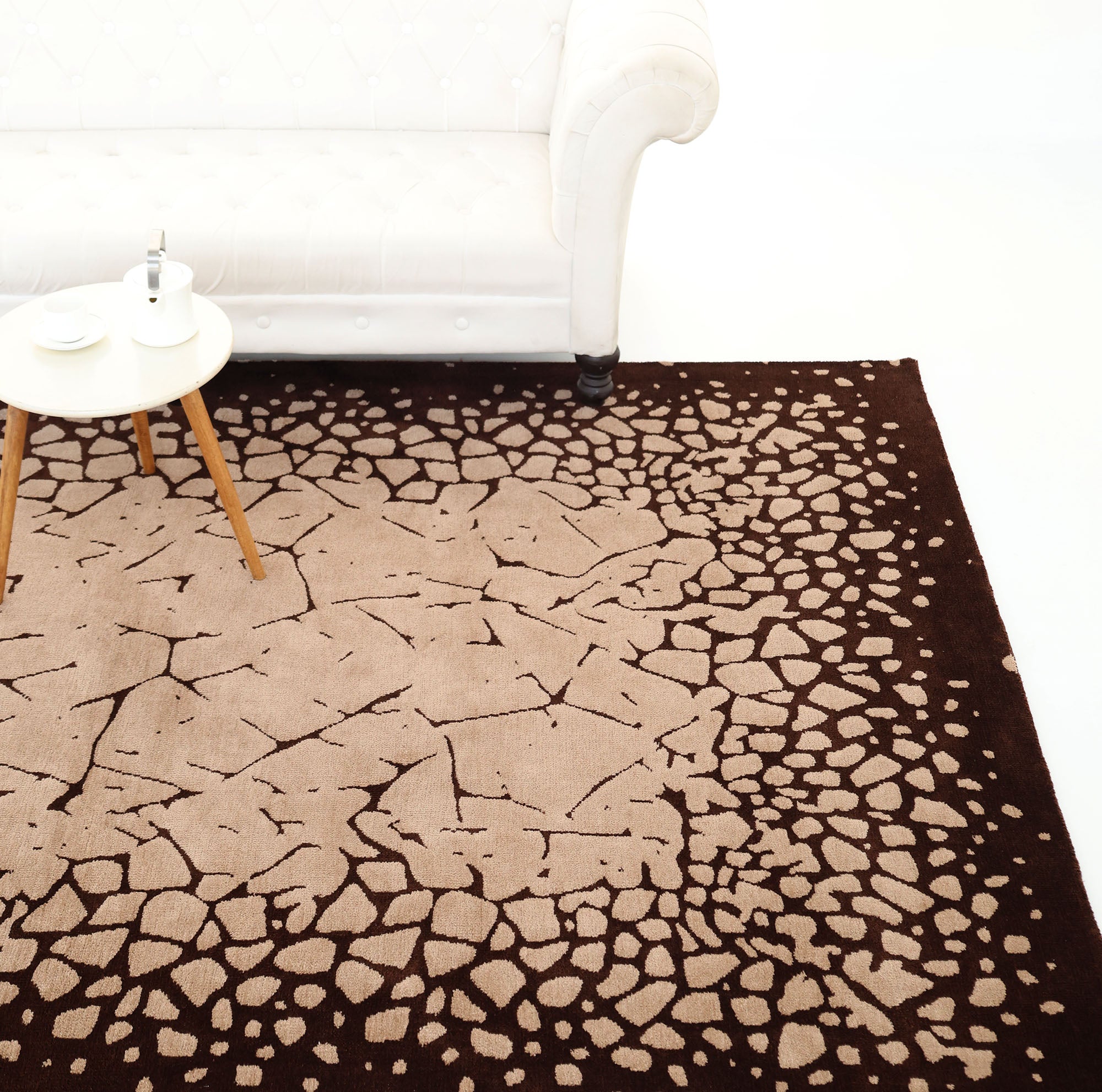 A modern living room with a white tufted sofa, a small round wooden table set with tea, and the RAMSHA HOME Terrain Echo Abstract Rug Shaped Rug CMN 118 in beige and dark brown, featuring a cracked, pebble-like pattern.