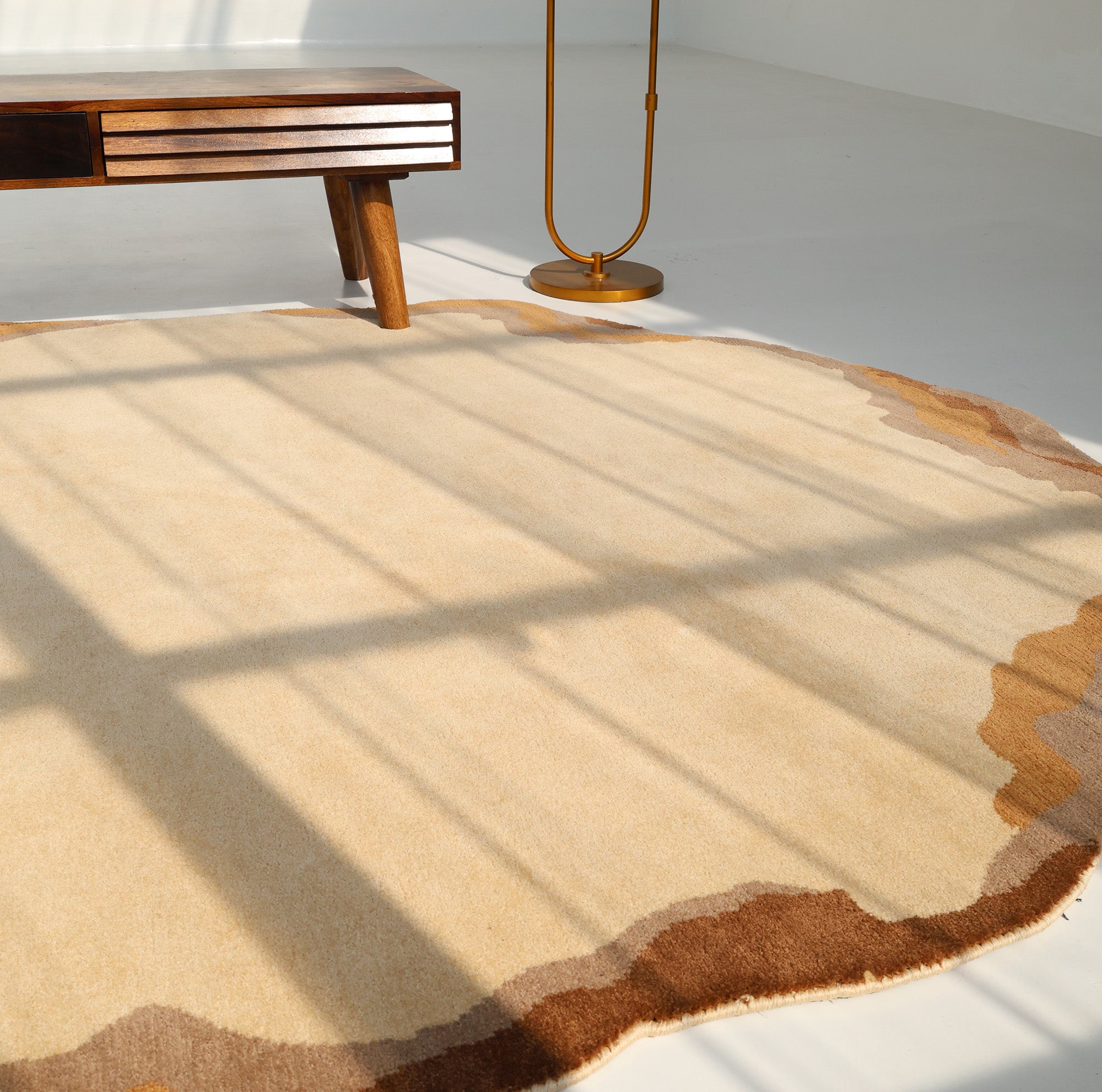 The Earth Layers Contemporary Rug Shaped Rug CMN 119 by RAMSHA HOME in beige with brown edges lies on a white floor, partly under a wooden table and next to a gold floor lamp, as sunlight casts window shadows across the peaceful space.