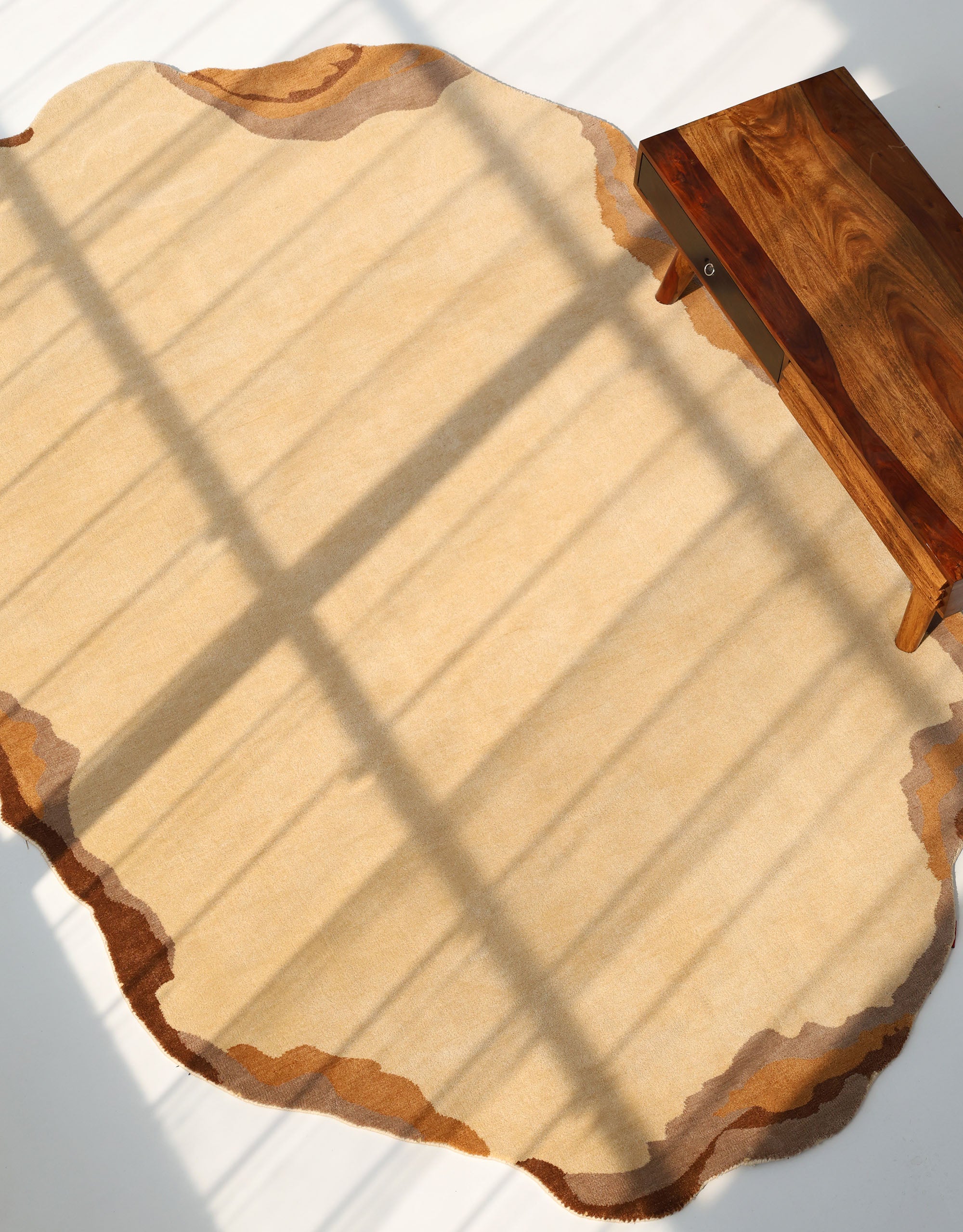 The RAMSHA HOME Earth Layers Contemporary Rug Shaped Rug CMN 119, featuring a beige base and irregular brown edges, lies partially beneath a wooden table as sunlight casts grid-like shadows on its abstract design.
