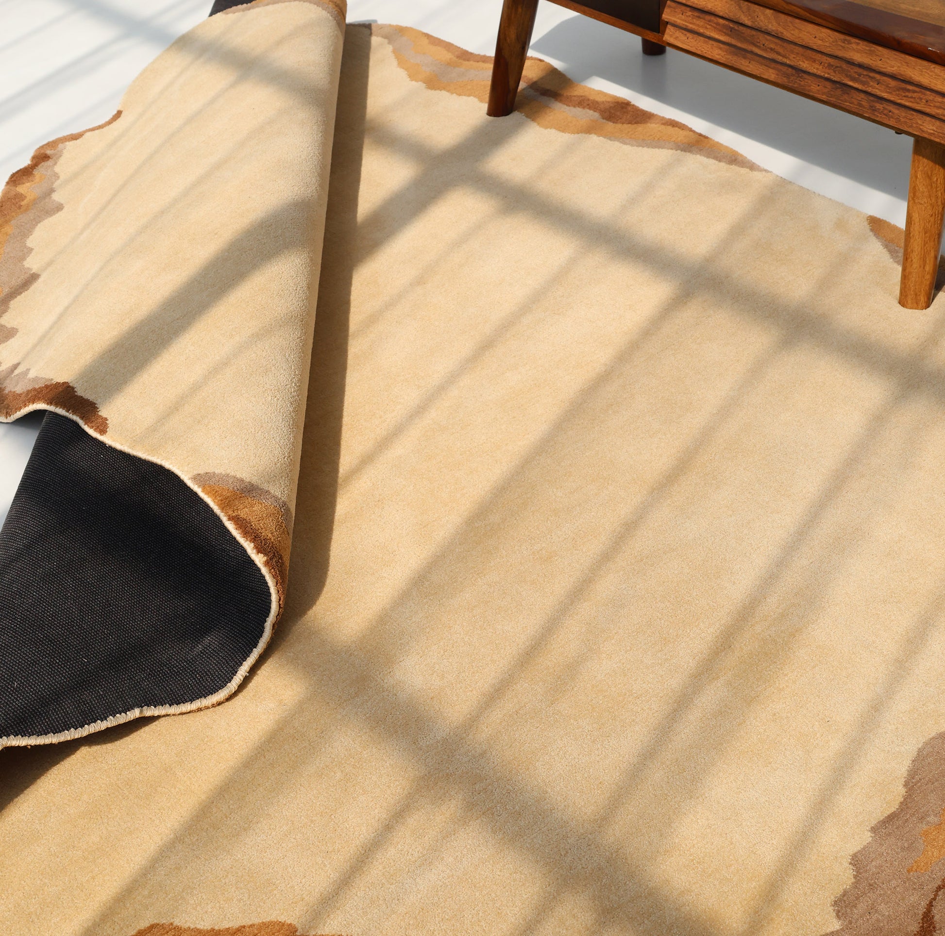 The RAMSHA HOME Earth Layers Contemporary Rug Shaped Rug CMN 119, featuring a beige abstract design with brown edges, is partially rolled on a light floor near a wooden chair as windowpane shadows stretch across the area.