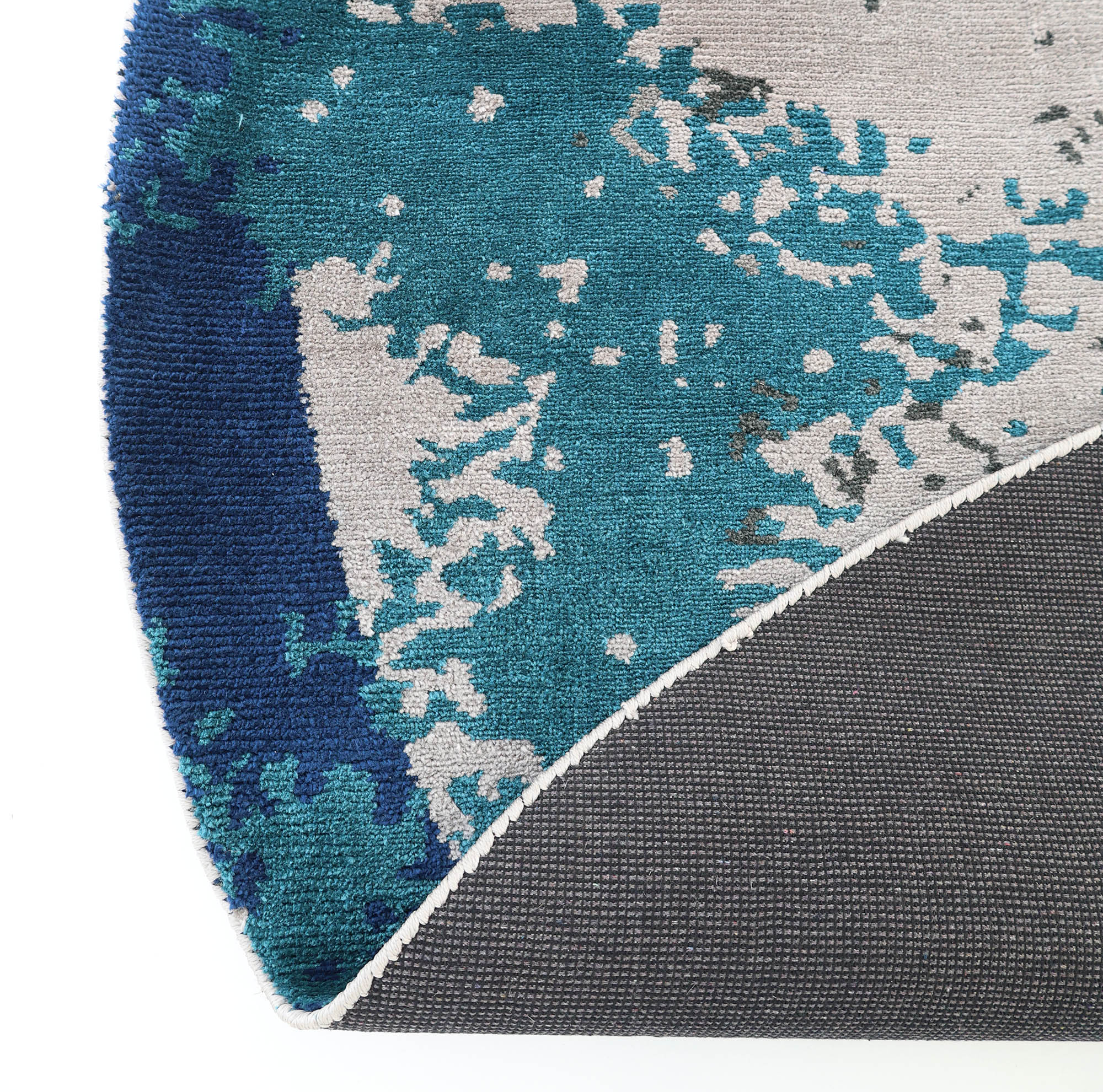 A close-up of the RAMSHA HOME Modern Blue Round Abstract Rug CMN 121 highlights its textured surface, abstract teal, blue, and gray geometric patterns, and non-slip black and white backing. The rug is slightly curled to show both sides.