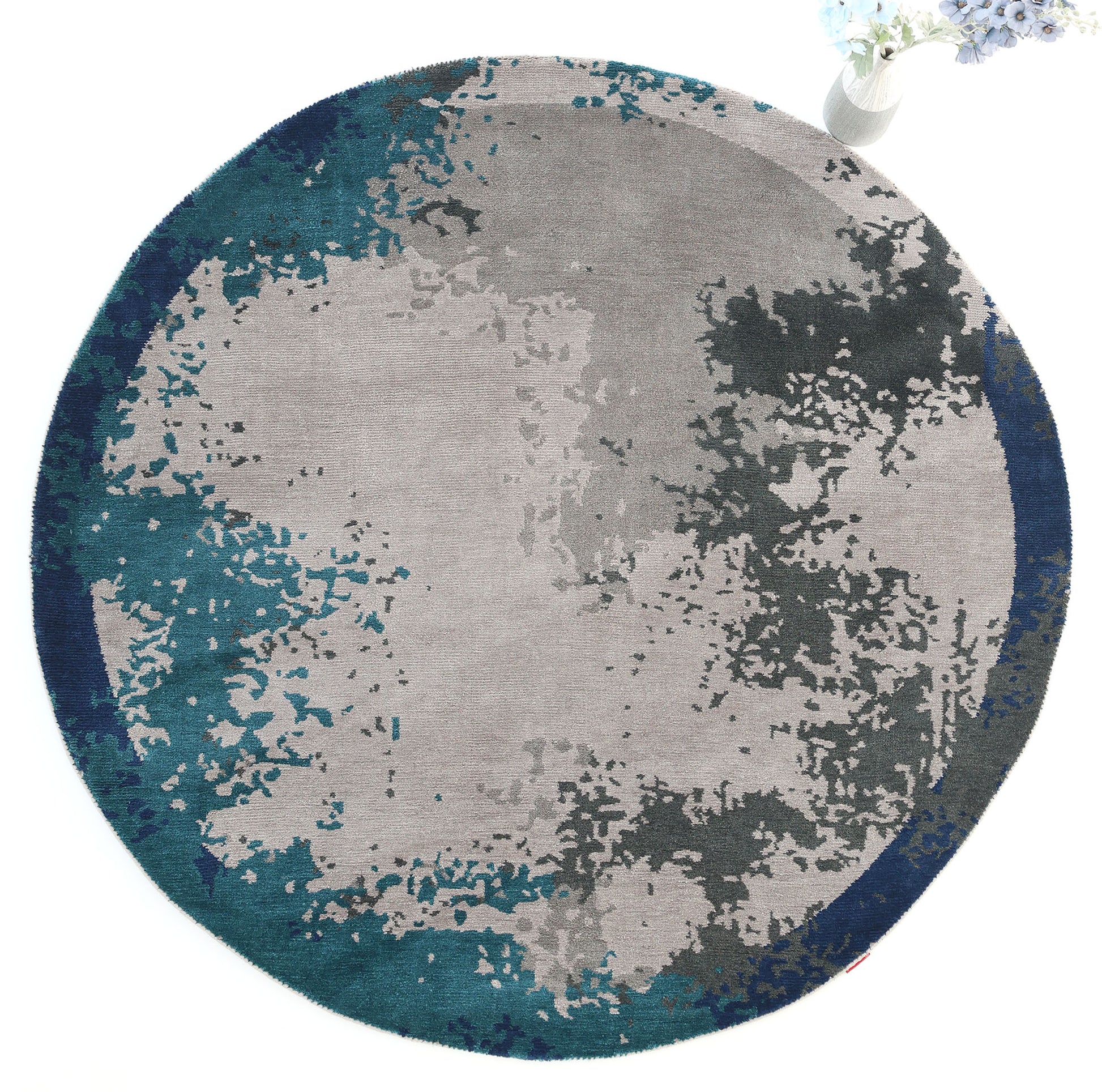 The RAMSHA HOME Modern Blue Round Abstract Rug CMN 121 showcases a textured, watercolor-inspired pattern in gray, teal, and dark blue. A vase with white flowers at the upper right edge enhances its contemporary appeal.