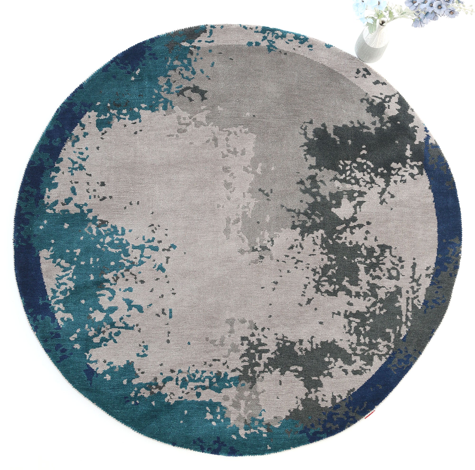 The RAMSHA HOME Modern Blue Round Abstract Rug CMN 121 showcases a textured, watercolor-inspired pattern in gray, teal, and dark blue. A vase with white flowers at the upper right edge enhances its contemporary appeal.