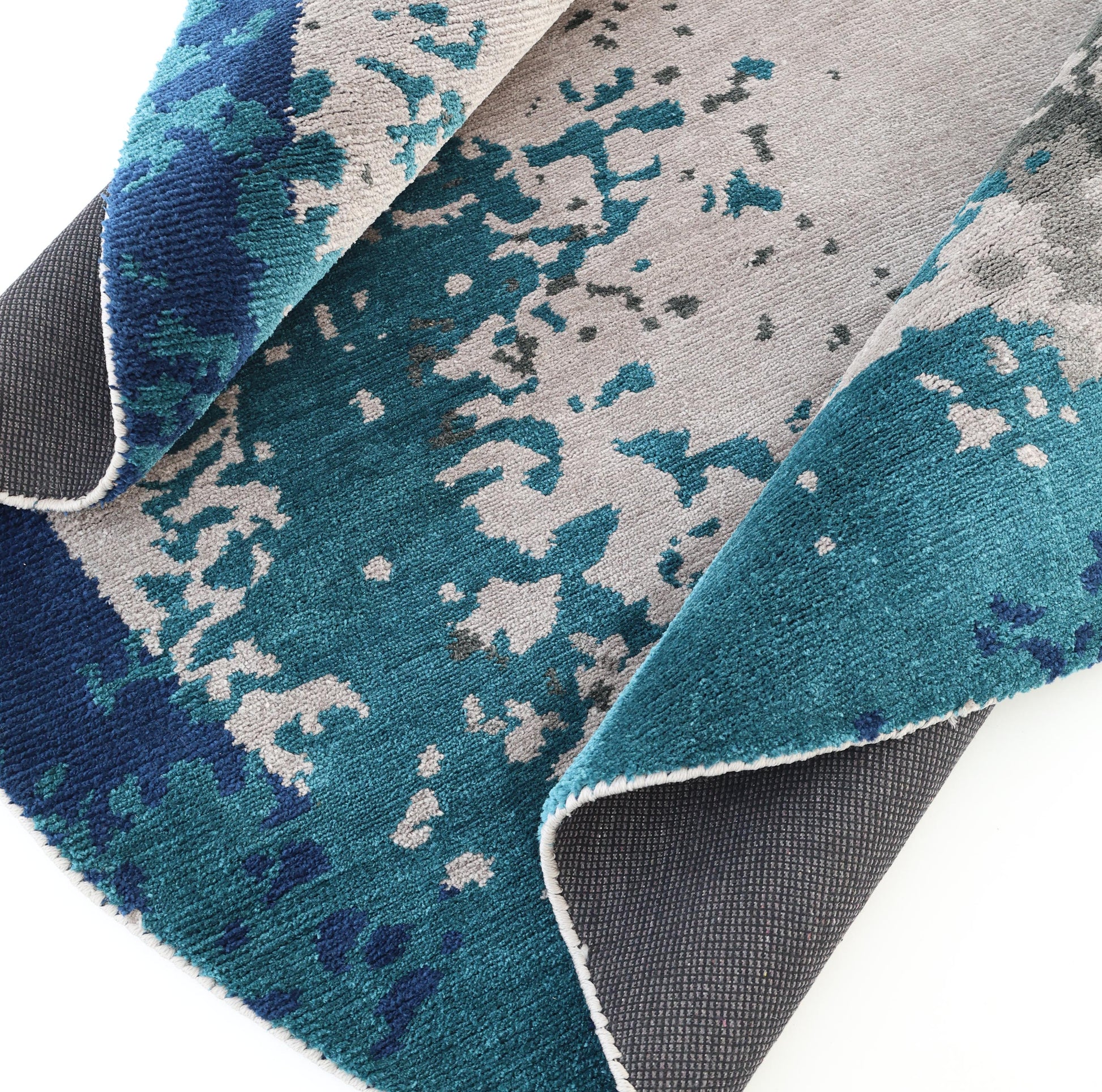 A close-up of the RAMSHA HOME Modern Blue Round Abstract Rug CMN 121 shows blue, teal, and gray abstract patterns, with its textured surface partially folded to reveal a dark gray underside.