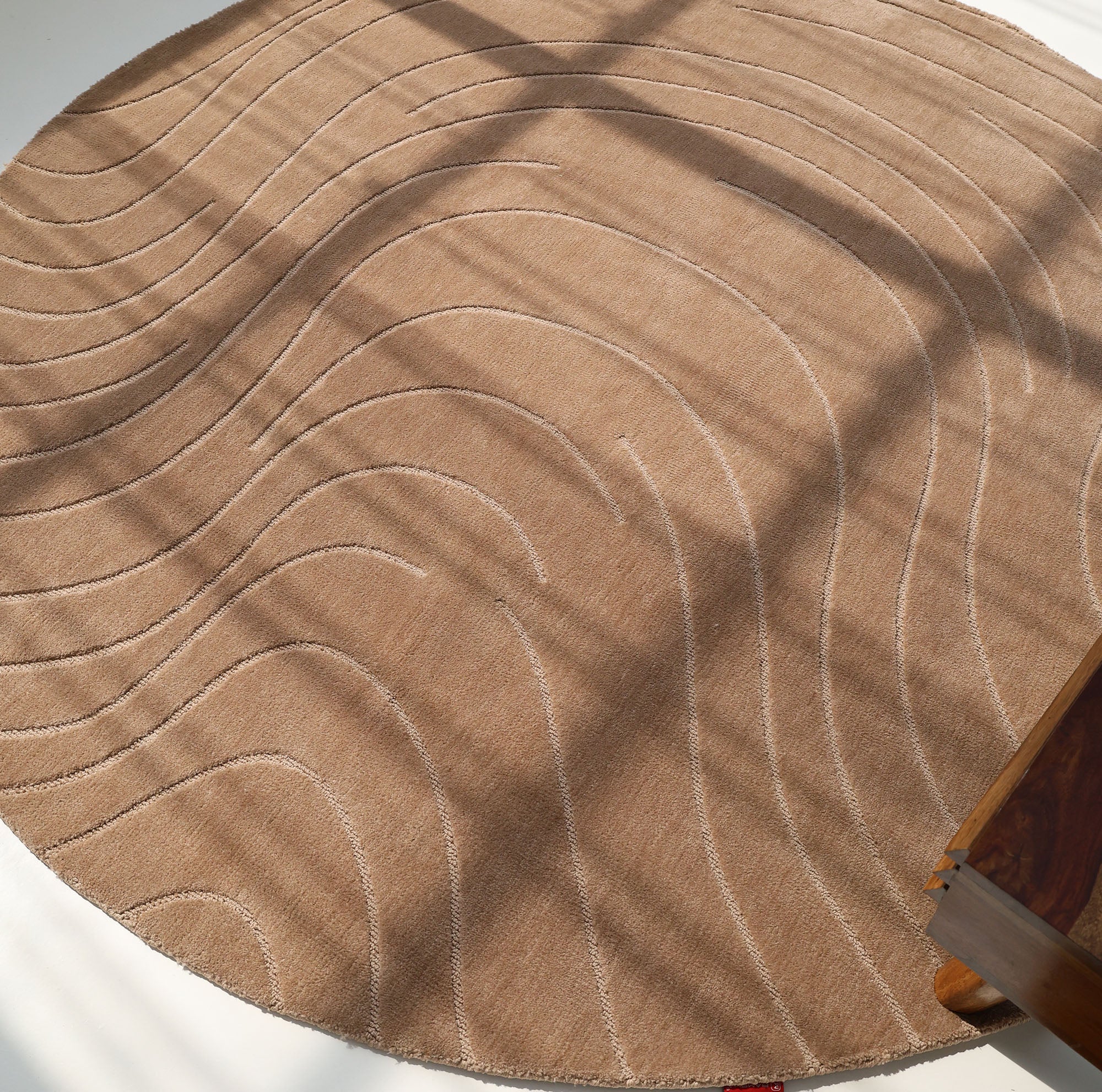 The RAMSHA HOME Modern Brown Round Abstract Rug CMN 124 features wavy cream lines in a striking pattern, with sunlight and shadows across its light brown surface and part of a wooden table visible at the right.