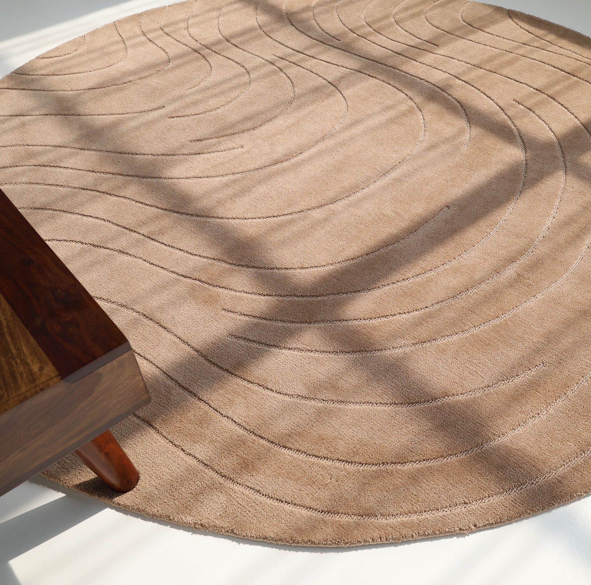 The RAMSHA HOME Modern Brown Round Abstract Rug CMN 124, with subtle curved lines and a beige hue, rests on a light floor bathed in sunlight. The lower left corner reveals part of a wooden table.