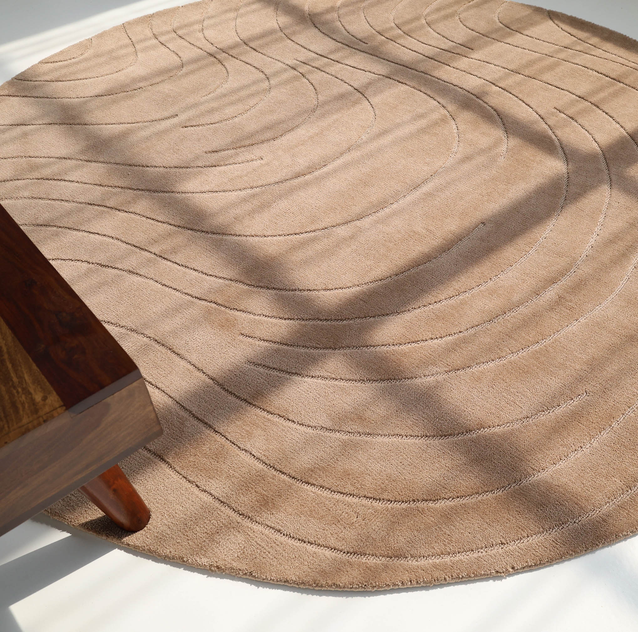 The RAMSHA HOME Modern Brown Round Abstract Rug CMN 124, with subtle curved lines and a beige hue, rests on a light floor bathed in sunlight. The lower left corner reveals part of a wooden table.