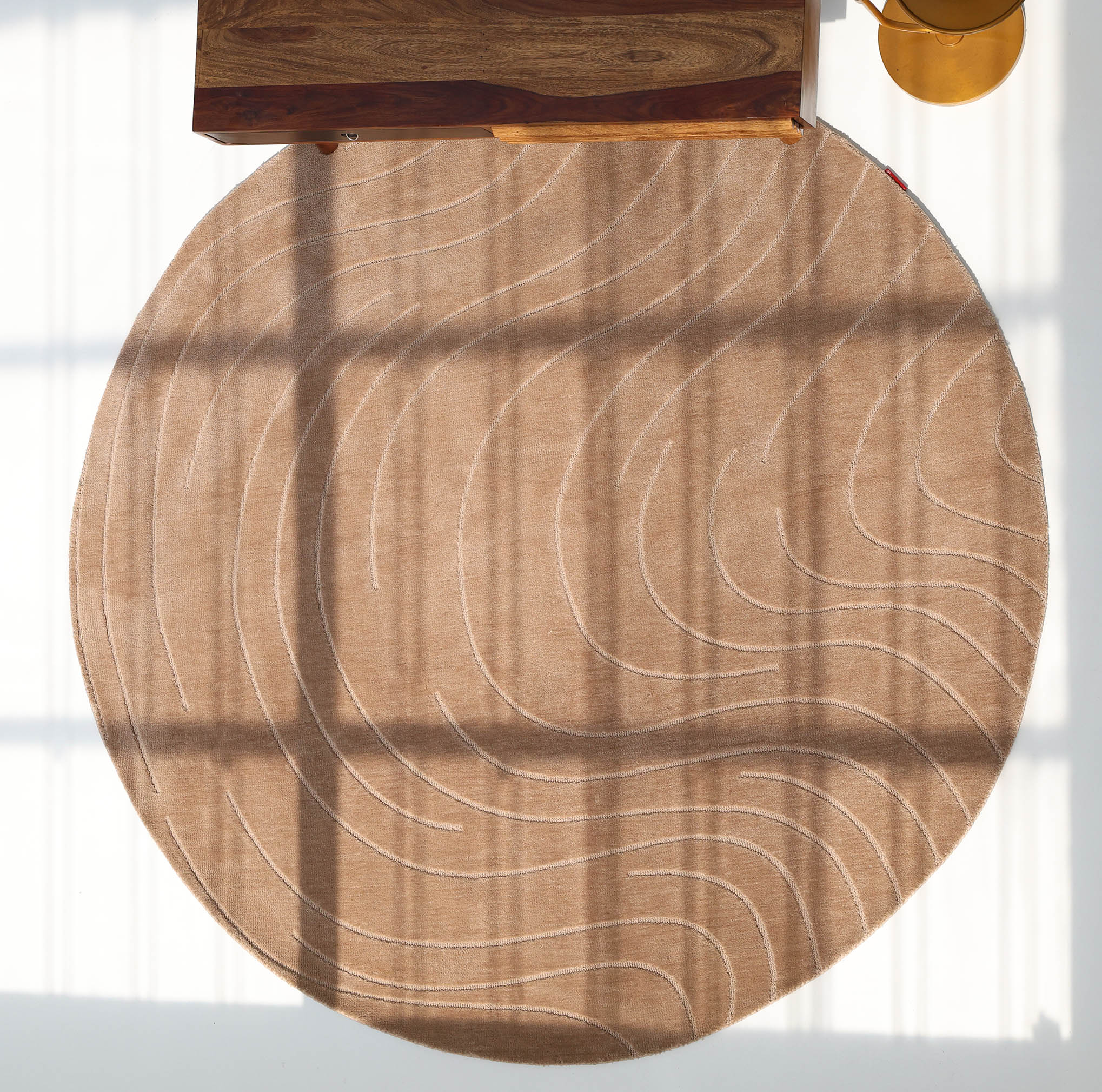 The RAMSHA HOME Modern Brown Round Abstract Rug CMN 124, featuring subtle wavy geometric patterns, lies on a white floor with sunlight casting windowpane shadows. A wooden table with a gold lamp appears at the edge of the contemporary scene.