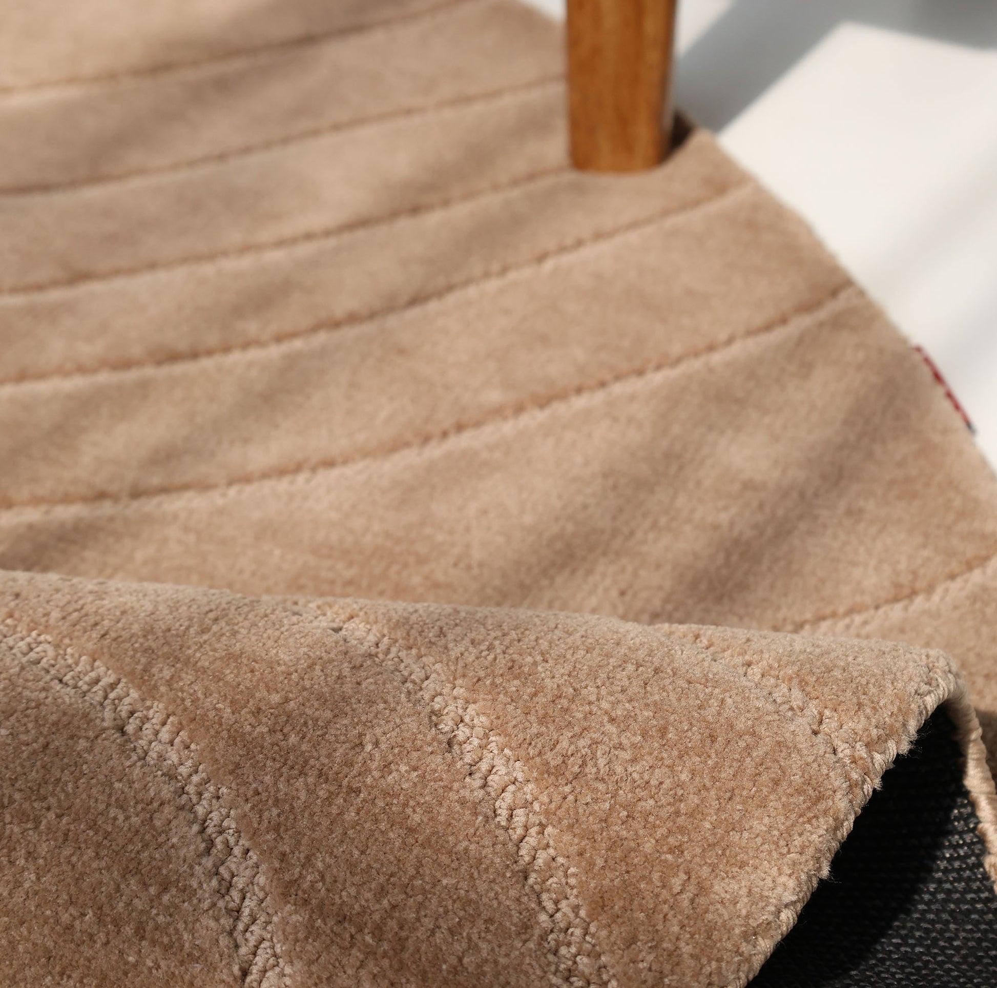 Close-up of the RAMSHA HOME Modern Brown Round Abstract Rug CMN 124: beige textured surface with stitched lines, black underside partially visible, and a wooden leg at the top. Sunlight casts soft shadows, highlighting its contemporary design.