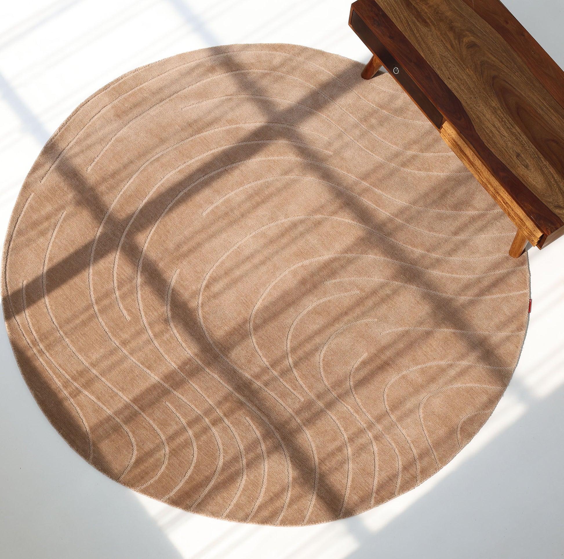 The RAMSHA HOME Modern Brown Round Abstract Rug CMN 124, featuring subtle wavy white lines, sits on a white floor beside a wooden bench as window shadows cast grid-like patterns across the rug and floor.