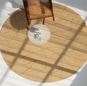 The Modern Sand Color Round Abstract Rug CMN 125 by RAMSHA HOME is shown from above with a small white table holding a teapot and cup, as a wooden chair casts a shadow across the contemporary geometric design.