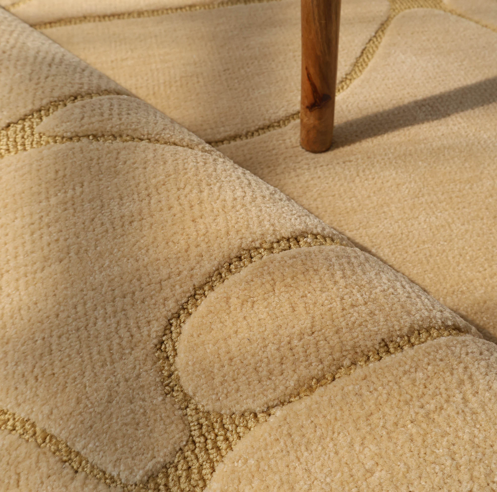 Close-up of the RAMSHA HOME Modern Sand Color Round Abstract Rug CMN 125, showcasing its raised curved and geometric patterns, with a wooden furniture leg casting a shadow on the textured surface.