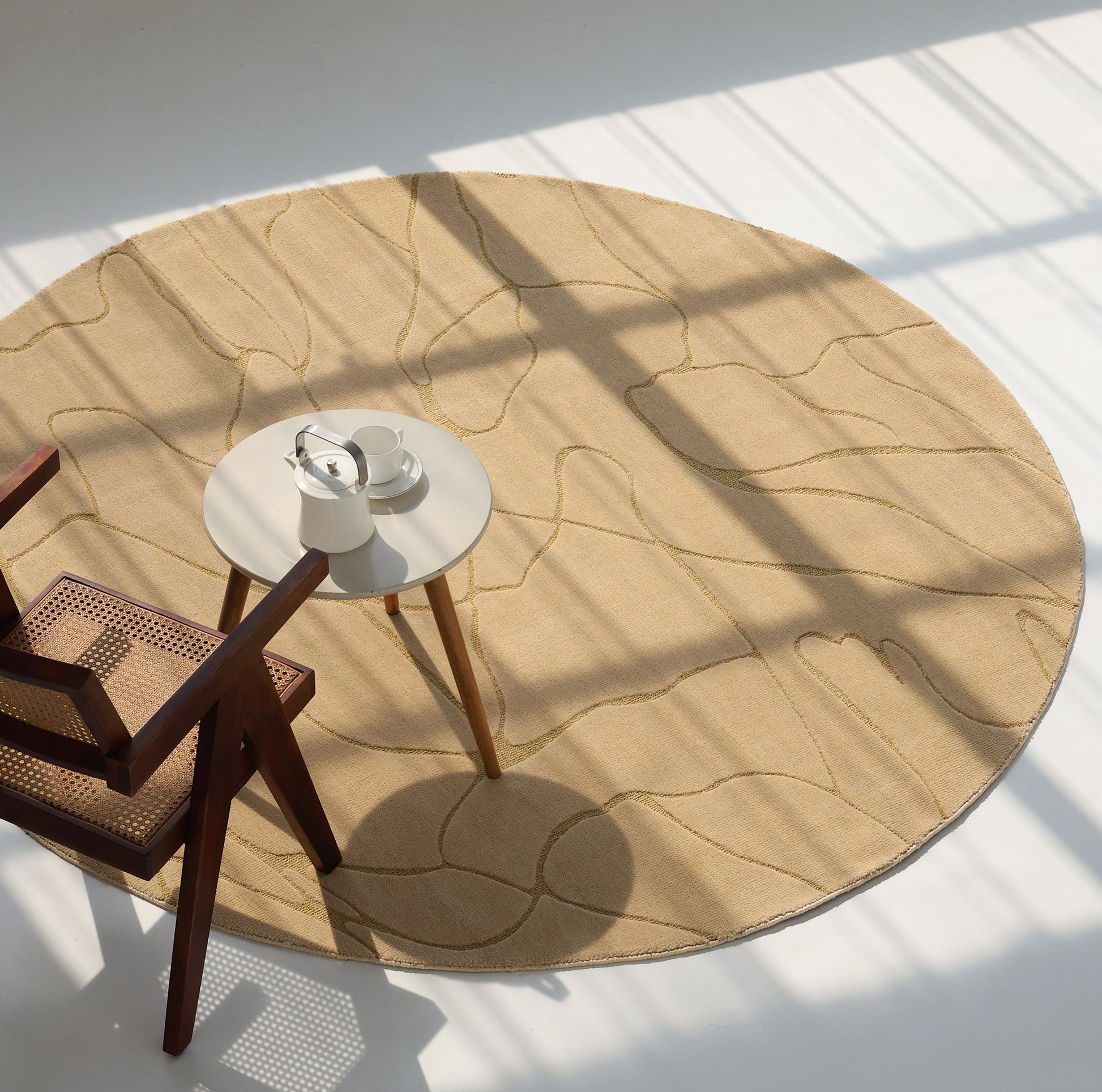 The Modern Sand Color Round Abstract Rug CMN 125 by RAMSHA HOME lies on the floor, holding a wooden chair with a woven seat and a small round table set with a teapot, cup, and saucer as sunlight streams through the window.