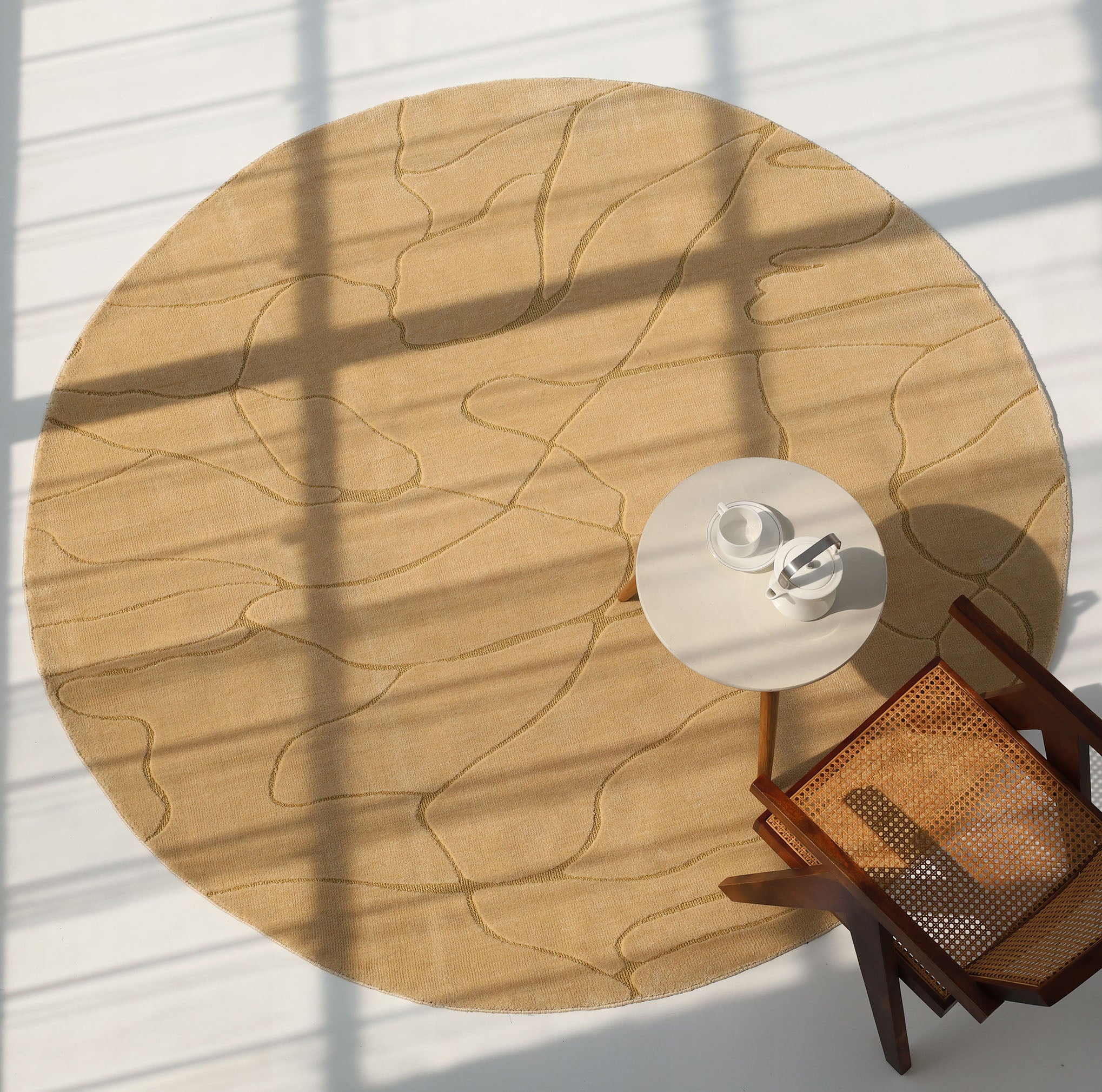 The Modern Sand Color Round Abstract Rug CMN 125 by RAMSHA HOME features subtle geometric patterns on a white floor, partially under a small round table with a cup and saucer. A wooden chair with woven seat is nearby, bathed in sunlight through window shadows.