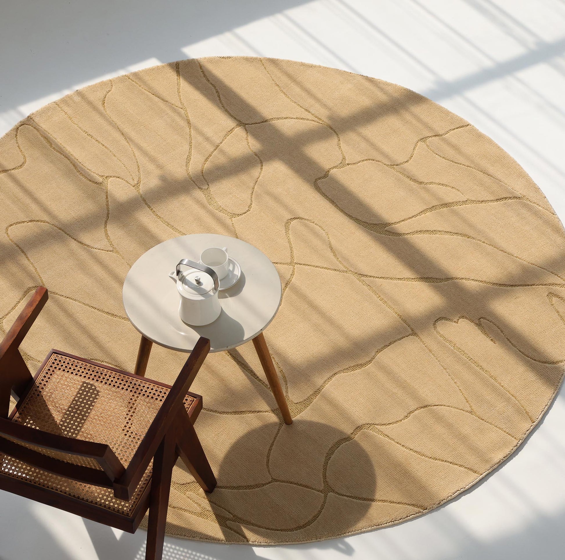 The Modern Sand Color Round Abstract Rug CMN 125 by RAMSHA HOME is bathed in sunlight, with a wooden chair with woven seat and a small round table set with a teapot and cups arranged on top.