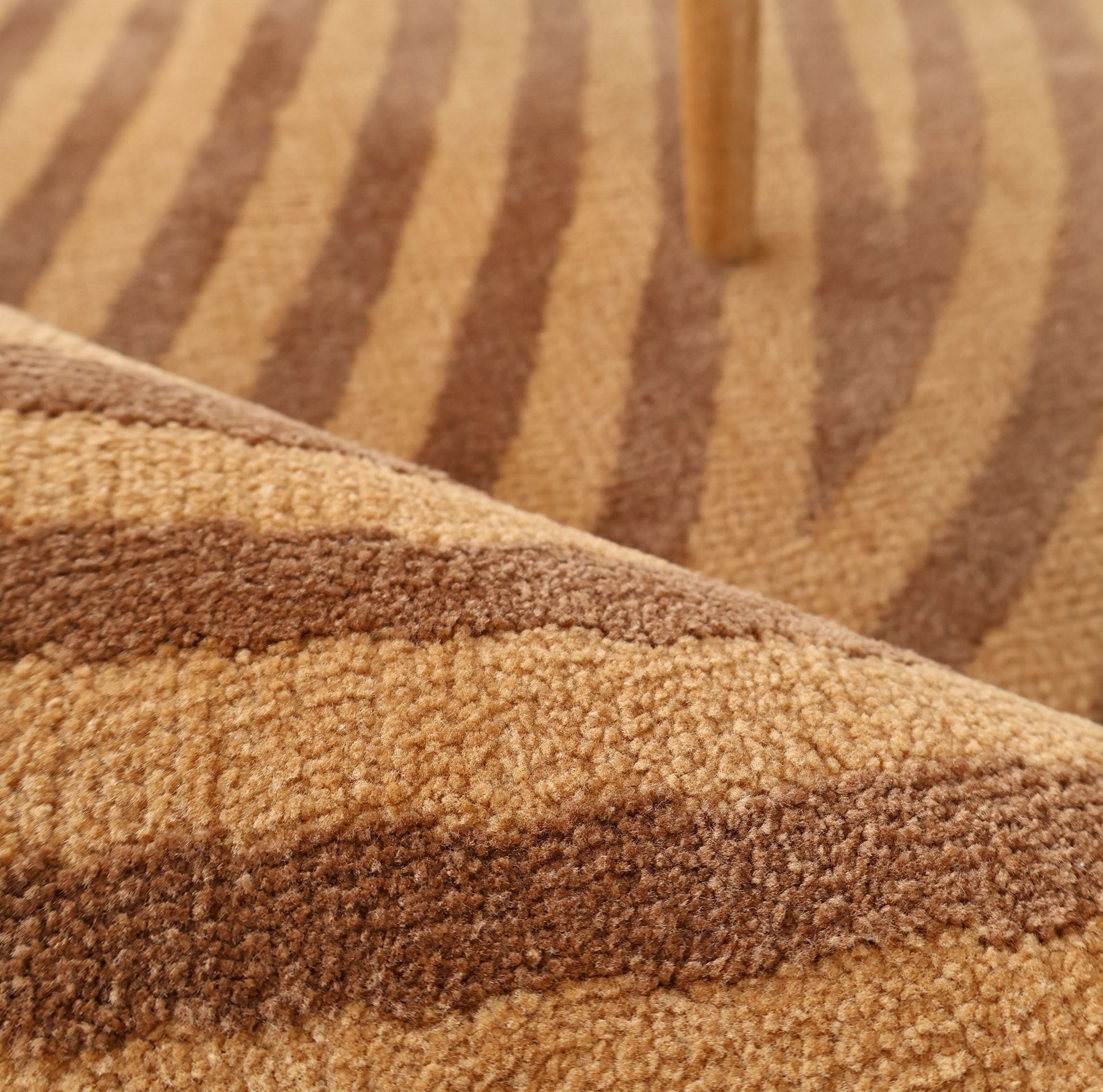 Close-up of the RAMSHA HOME Modern Brown Color Round Abstract Rug CMN 130 featuring beige and brown geometric patterns, a rolled edge in the foreground, and a wooden furniture leg in the background.
