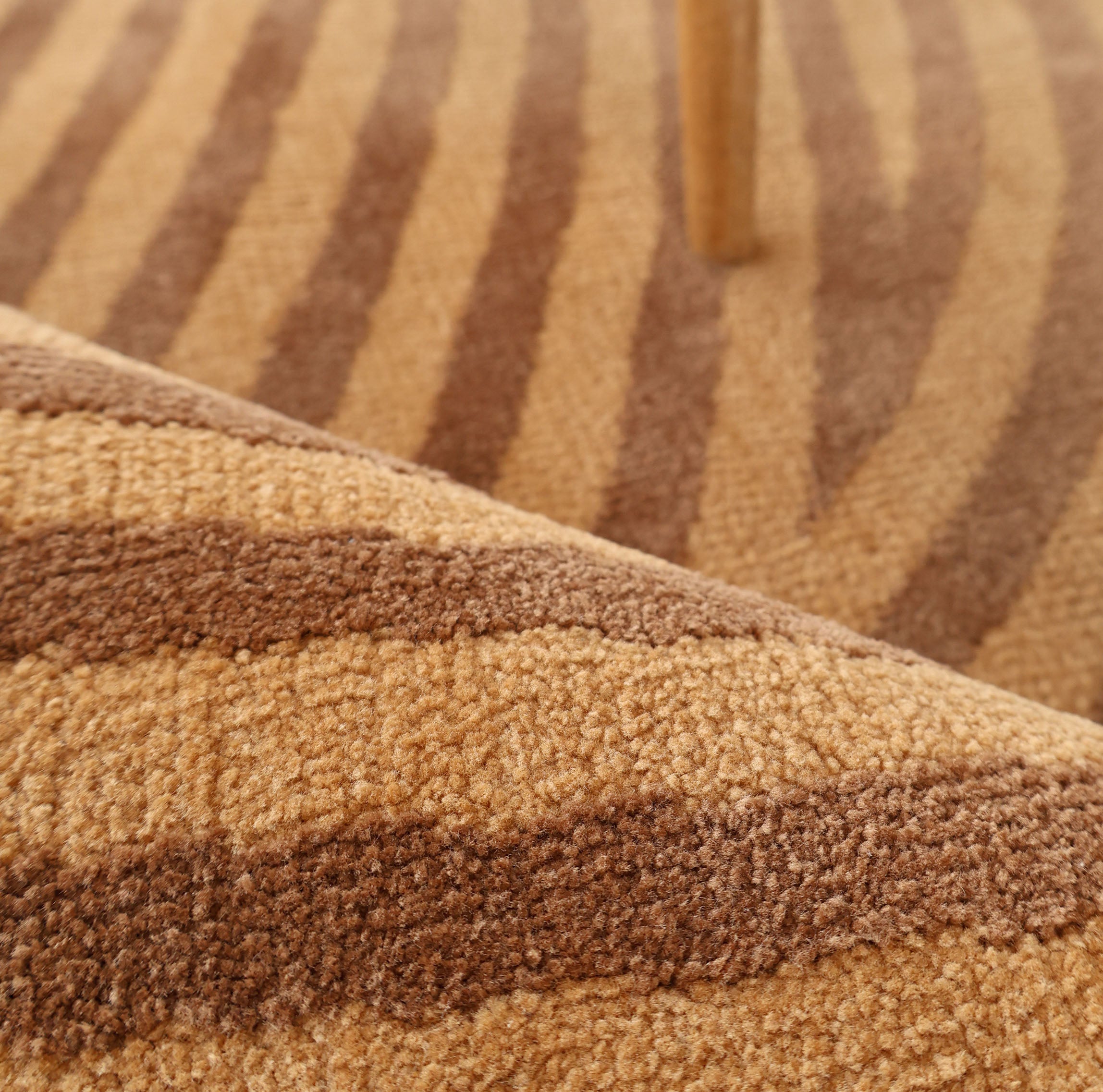 Close-up of the RAMSHA HOME Modern Brown Color Round Abstract Rug CMN 130 featuring beige and brown geometric patterns, a rolled edge in the foreground, and a wooden furniture leg in the background.