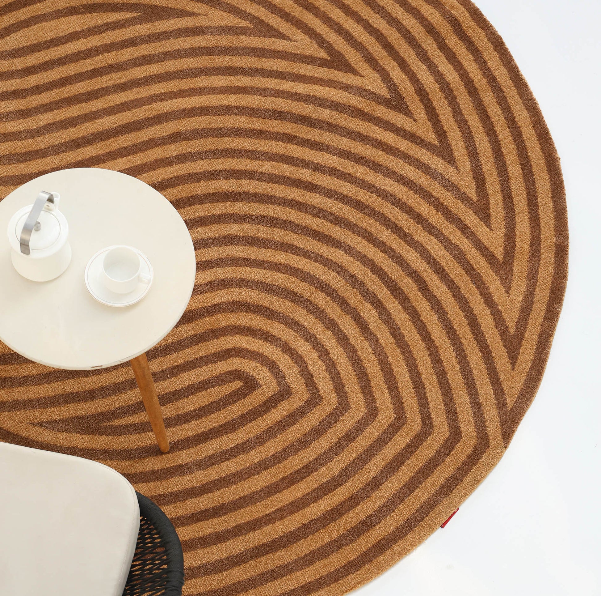 The RAMSHA HOME Modern Brown Color Round Abstract Rug CMN 130, featuring brown and tan geometric patterns, is placed beside a small round white table with a coffee pot and cup; part of a white chair appears nearby.