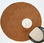 The RAMSHA HOME Modern Brown Color Round Abstract Rug CMN 130 with wavy stripes is shown from above, paired with a small white table holding a teapot and two cups, and part of a modern chair with a light cushion.