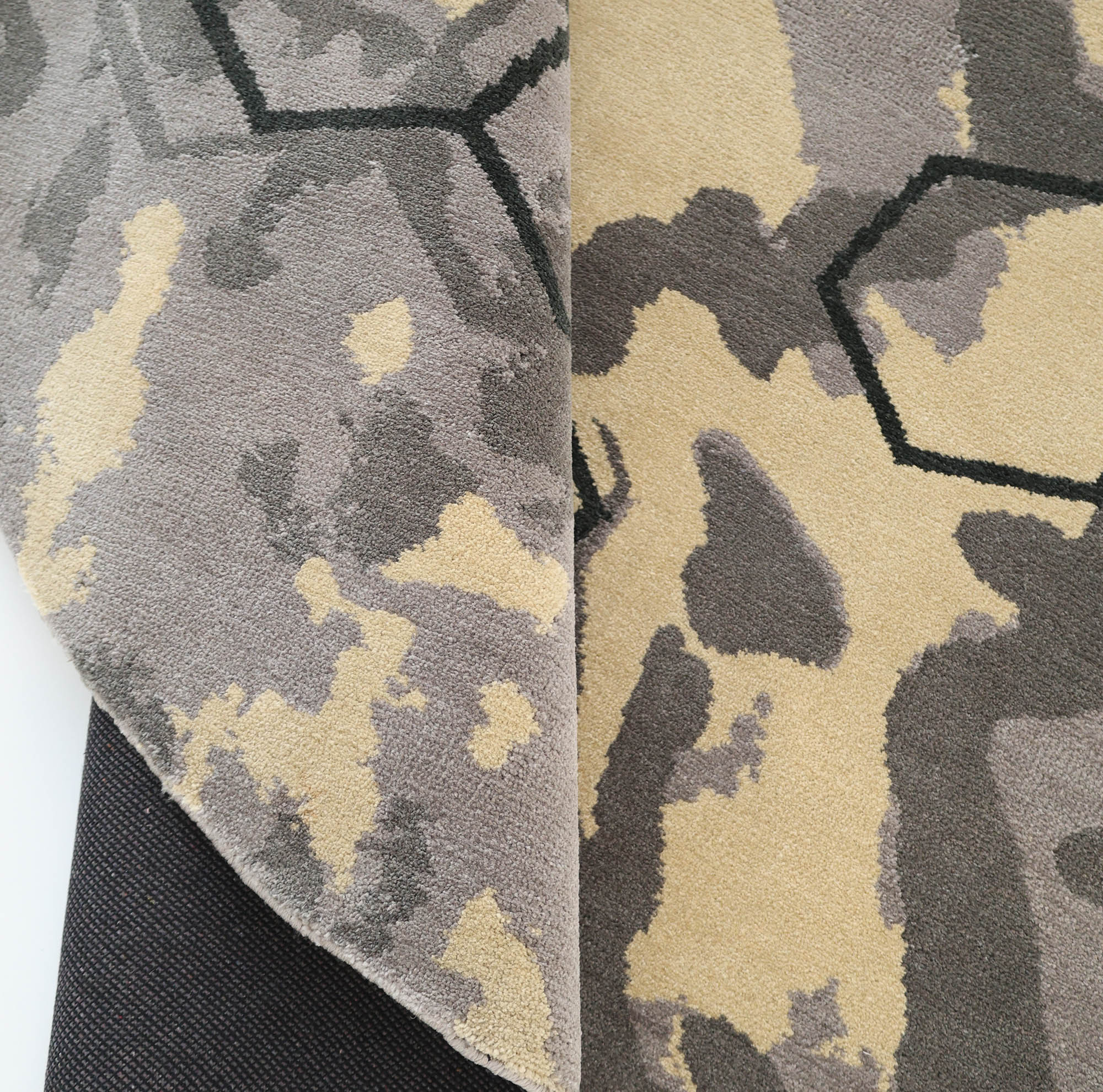 A close-up of the RAMSHA HOME Modern Geometric Round Rug CMN 133 in grey and beige, partially folded to show its underside and a black non-slip rug pad underneath.
