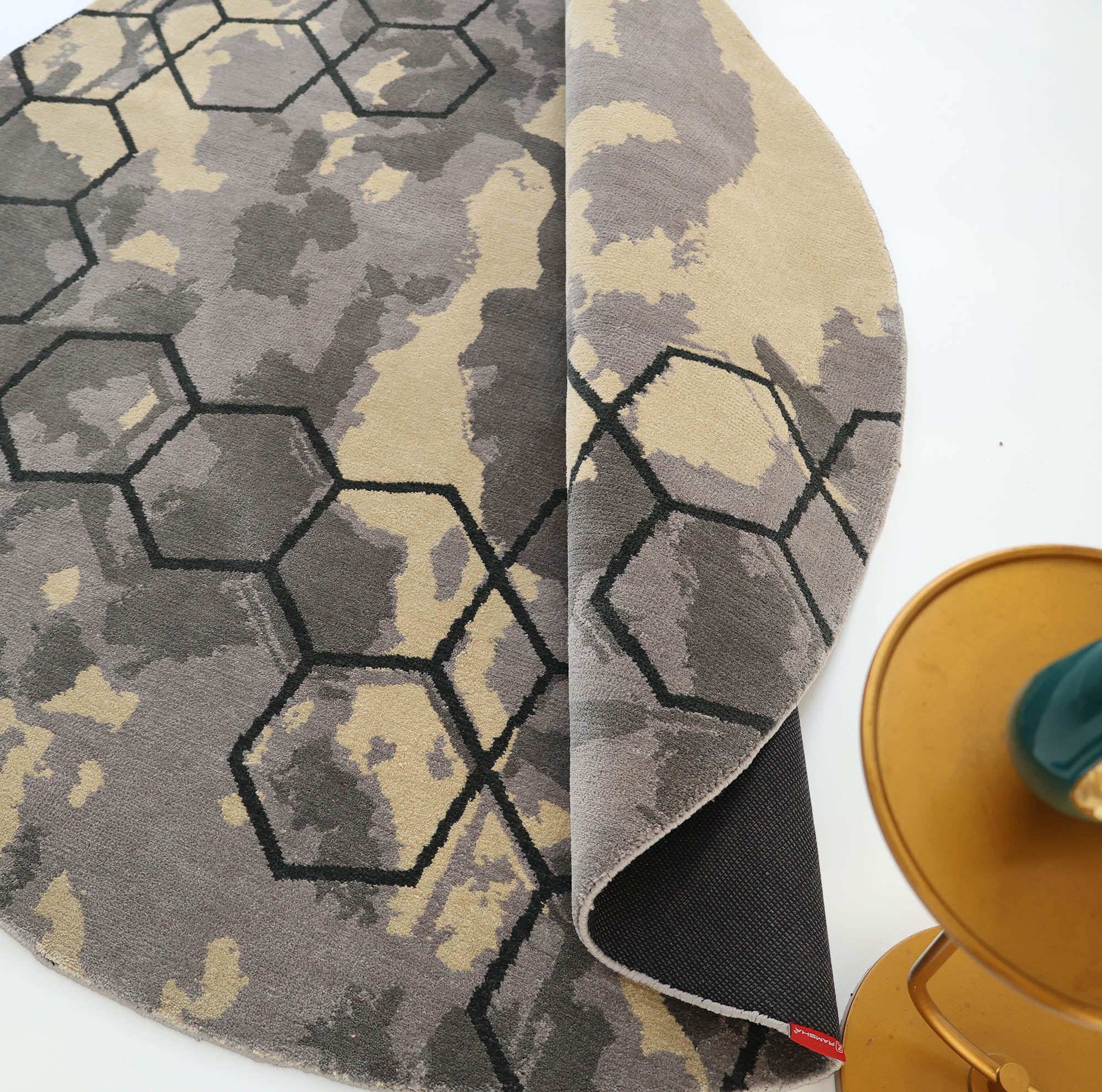 The RAMSHA HOME Modern Geometric Round Rug Grey & Beige Rug CMN 133 features a contemporary hexagonal pattern in beige, gray, and black. Shown partially flipped, it complements a golden side table nearby.