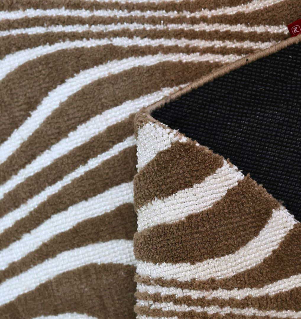 A close-up of the Oval Shape Novara Carpet Multi Color CMN-17 by RAMSHA HOME shows its brown and white zebra-striped design with one corner folded over, revealing a black non-slip backing underneath.