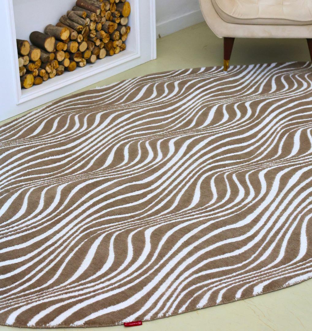 The Oval Shape Novara Carpet Multi Color CMN-17 by RAMSHA HOME features modern abstract wavy brown and white stripes, adding style beside firewood and a beige armchair with wooden legs.