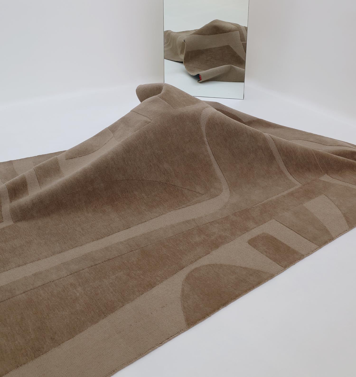 The RAMSHA HOME Dark Tone Multi Color Polyester CMN-19 rug drapes over a hidden object, forming a hill-like shape. A mirror behind the rug reflects the minimalistic room’s white walls and reveals part of a red object beneath it.