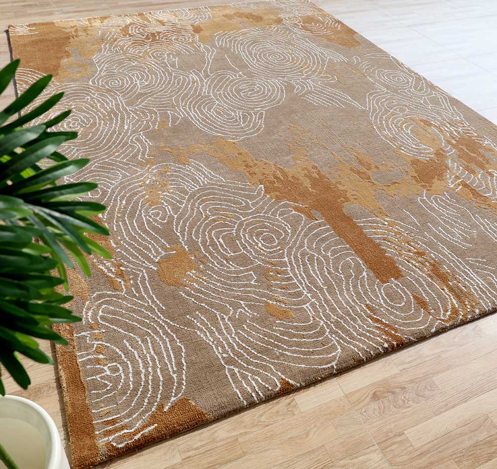 The RAMSHA HOME Multi Color Novara Decor Rug (CMN 33, 205x310cm) displays abstract swirls in beige, brown, and gold with white accents. Shown on a light wood floor with a green leafy plant nearby.