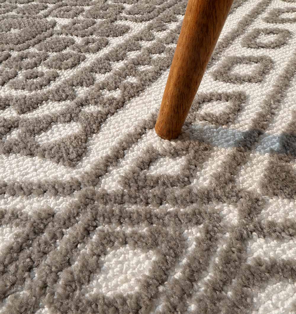 Close-up of a wooden furniture leg on the RAMSHA HOME Novara Rug - CMN 54 (180x280cm), featuring a grey and white diamond pattern. Sunlight casts a soft shadow across the machine-made surface.