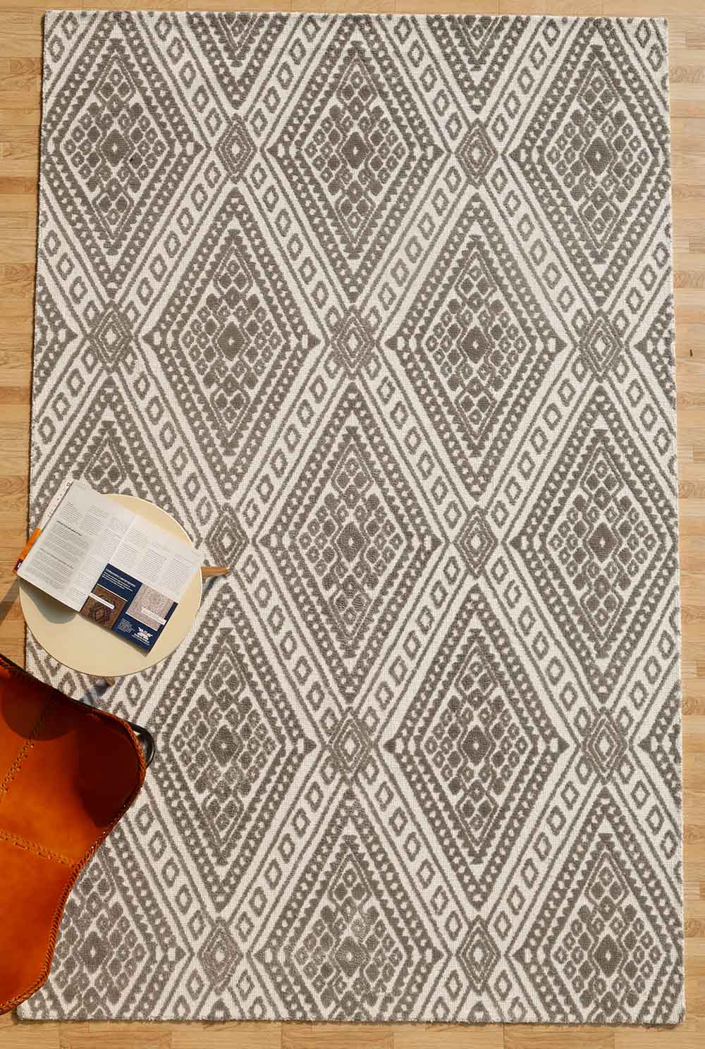 The RAMSHA HOME Grey & White Novara Rug - CMN 54 (180x280cm) features a geometric diamond pattern, displayed on a wooden floor with a round table, open magazine, and brown leather chair, adding modern elegance to the room.