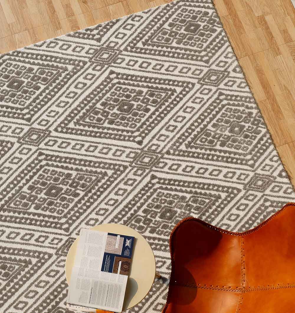 The RAMSHA HOME Grey & White Color Novara Rug - CMN 54 (180x280cm), featuring a geometric diamond pattern, is spread over a wooden floor beside a round beige table with an open book and a brown leather chair in the corner.