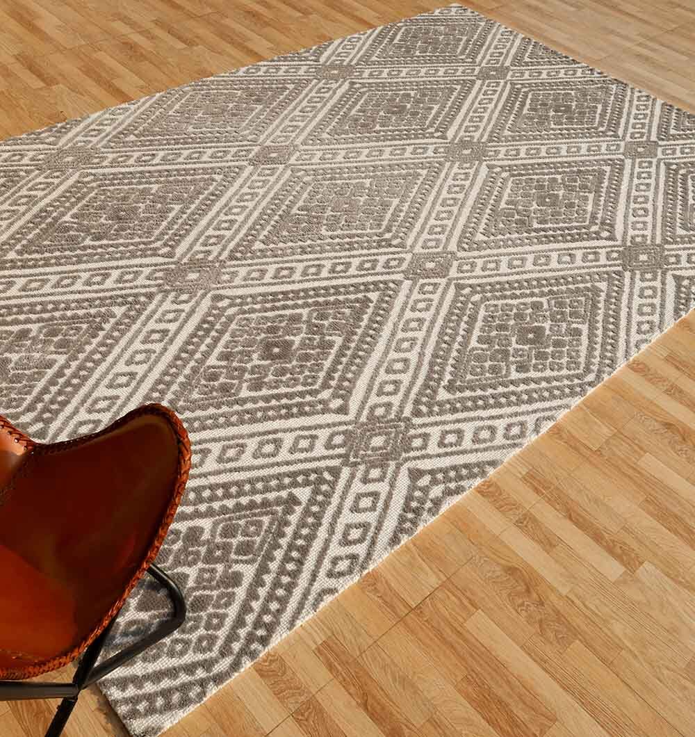 The RAMSHA HOME Grey & White Color Home Decor Novara Rug - CMN 54 (180x280cm) with a geometric diamond pattern lies on a light wood floor, next to a brown leather chair visible in the lower left corner.