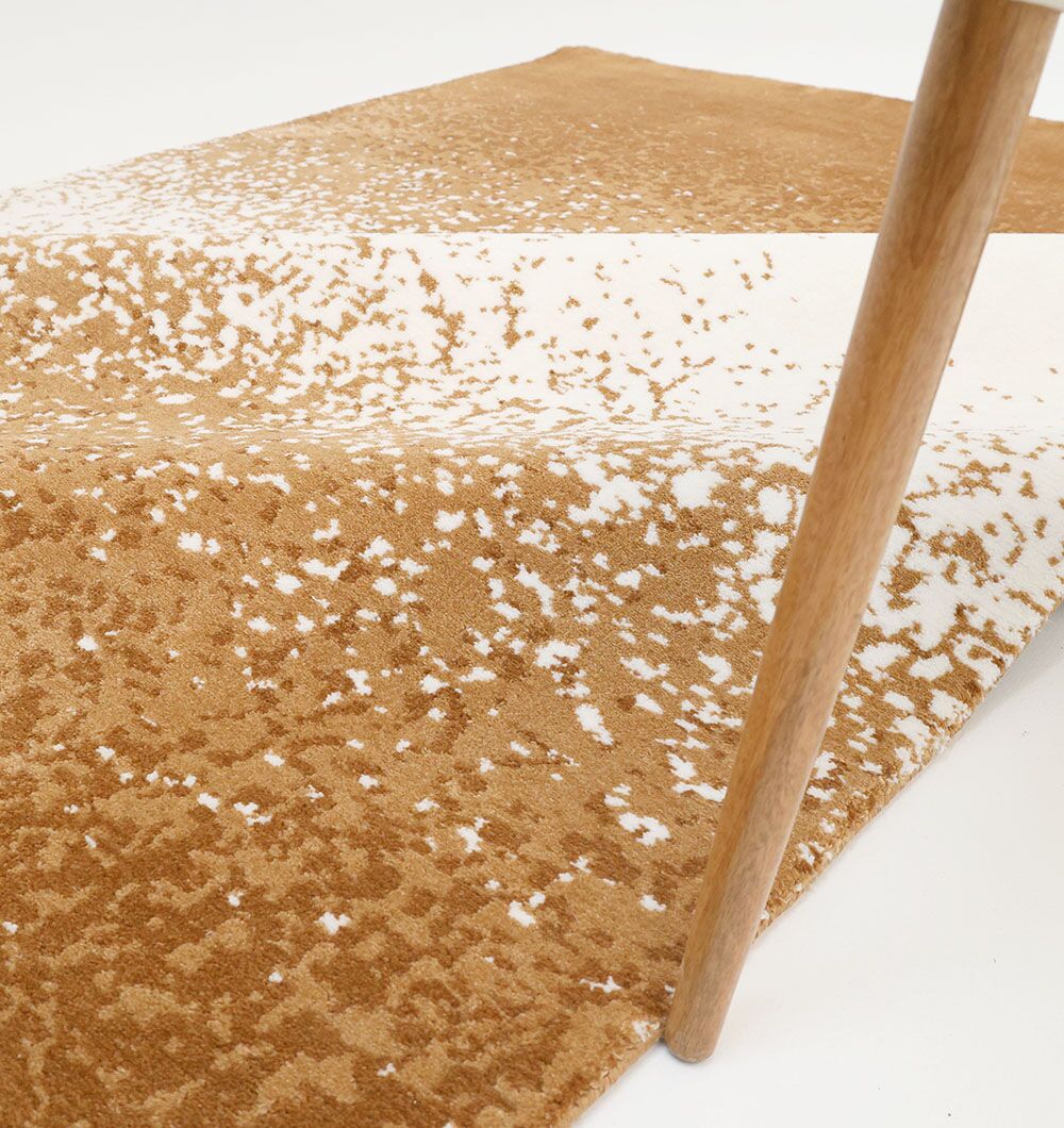 A close-up of the RAMSHA HOME Rectangle Multi Color Rug - CMN 69 (200X300cm) under a wooden furniture leg highlights its soft texture and modern pattern in tan and white tones.