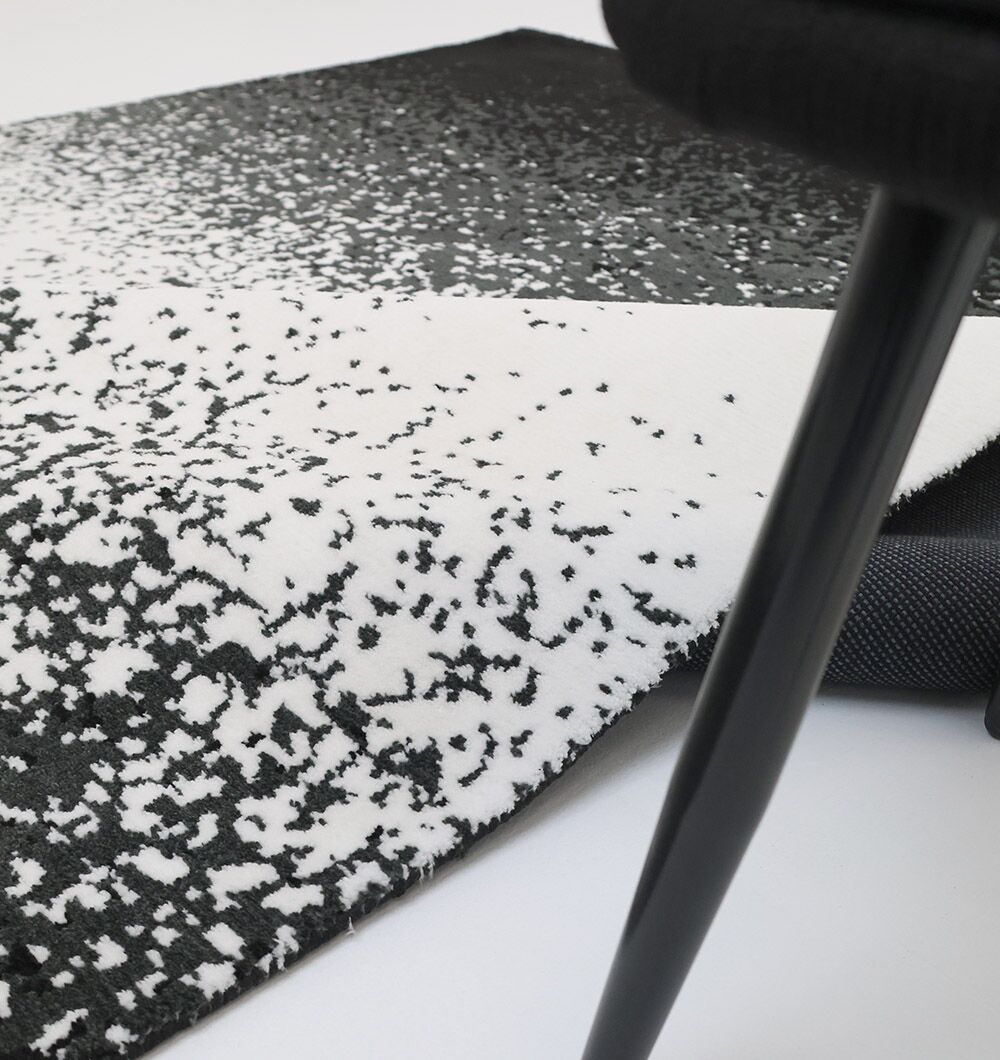 A close-up of the RAMSHA HOME Rectangle Multi Color Rug - CMN 71 (200x300cm) with a corner folded, shown beside a black chair leg on a light floor.