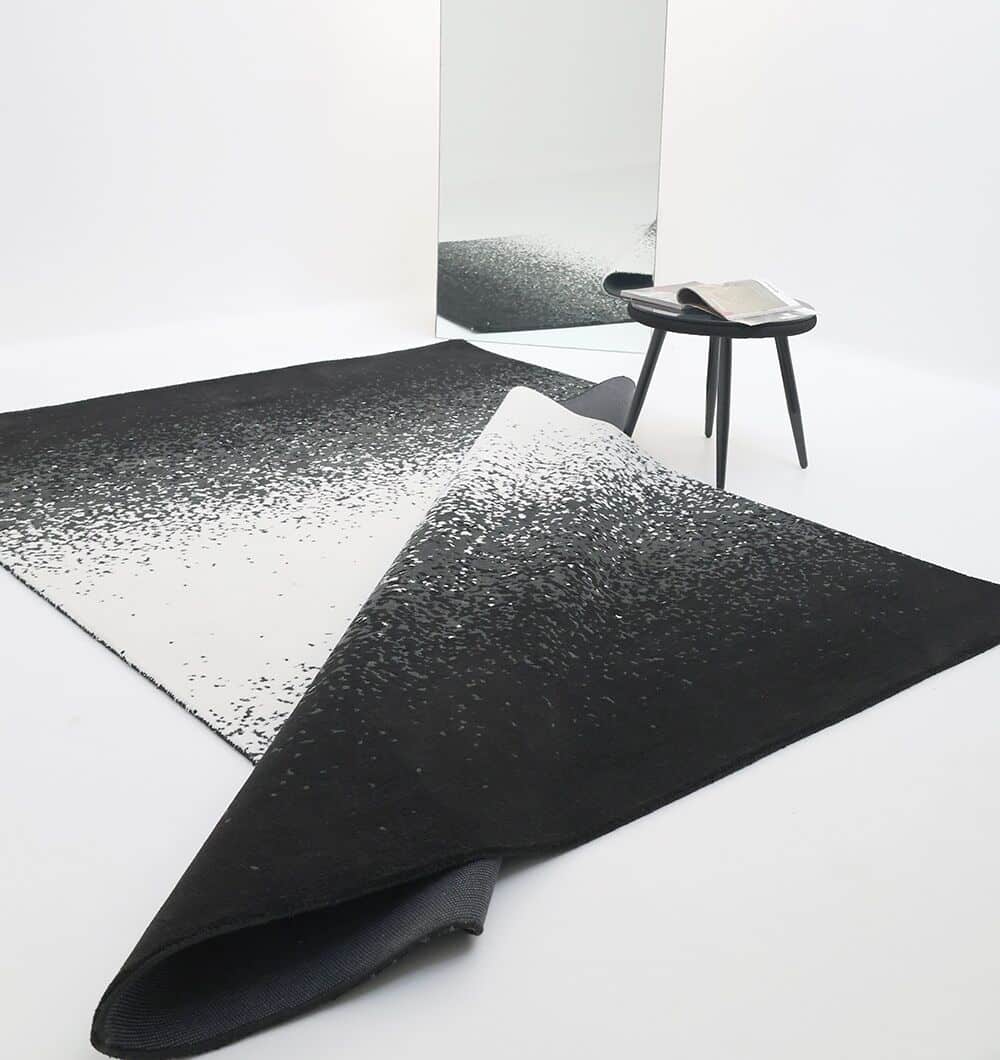 A modern room features the RAMSHA HOME Rectangle Multi Color Rug - CMN 71 (200x300cm), partly flipped, a small round black side table with a book, and a large rectangular mirror leaning against a white wall.