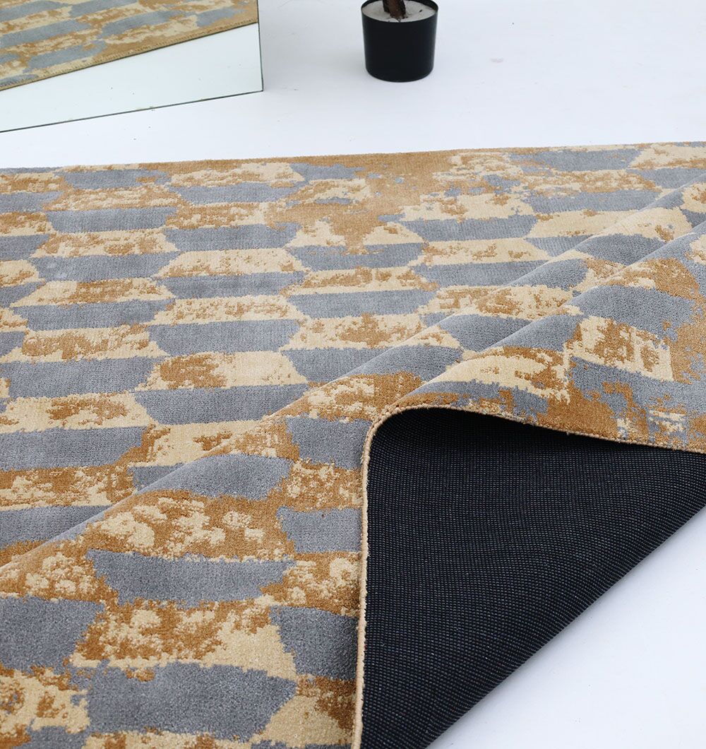 A close-up of the RAMSHA HOME Home Decor Multi Color Novara Rug - CMN 82 shows its geometric gold and gray pattern, with one corner folded back to reveal a black non-slip backing. A mirror and small potted plant are partially visible in the background.