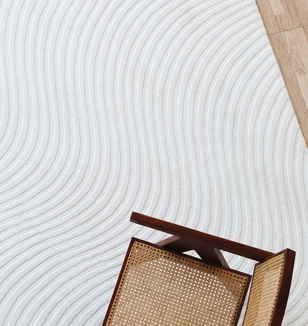 A wooden chair with a woven cane seat sits on the Rectangle White Color Novara Rug - CMN 85 (200x300 cm) by RAMSHA HOME, its white surface featuring curved wave patterns. Part of a wooden floor appears in the top right corner.