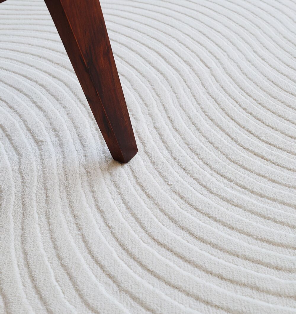 A dark wooden furniture leg rests on the Rectangle White Color Novara Rug - CMN 85 (200x300 cm) by RAMSHA HOME, its evenly spaced, wavy lines bringing a subtle modern pattern to this neutral rug.