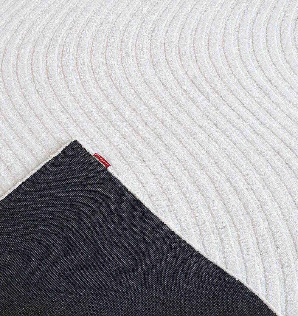 A close-up of the RAMSHA HOME Rectangle White Color Novara Rug - CMN 85 (200x300 cm) shows wavy parallel lines on its white textured surface, with a black corner folded to reveal a small red tag.