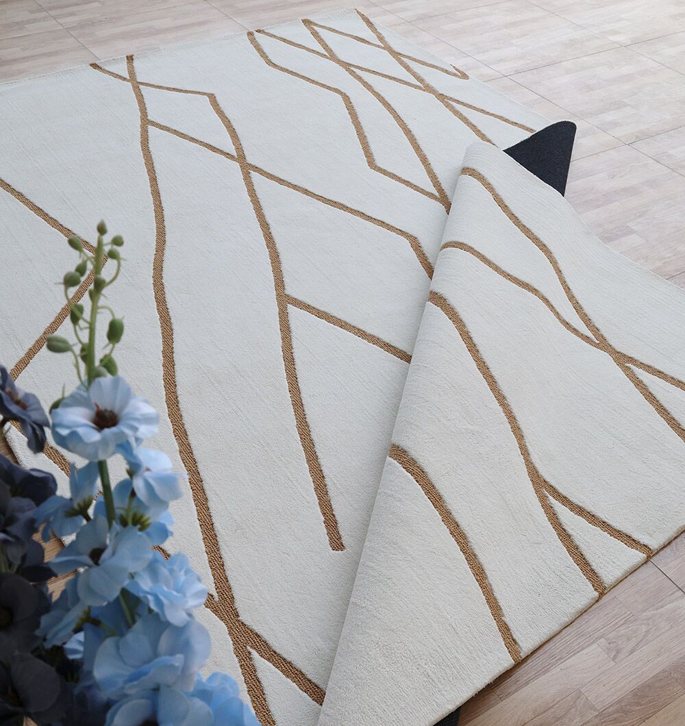 The RAMSHA HOME Home Decor Novara Area Rug - CMN 87 (200x300 cm) features a beige background with brown abstract lines, blue floral accents, and a non-slip black backing, displayed partially flipped on a light wood floor.