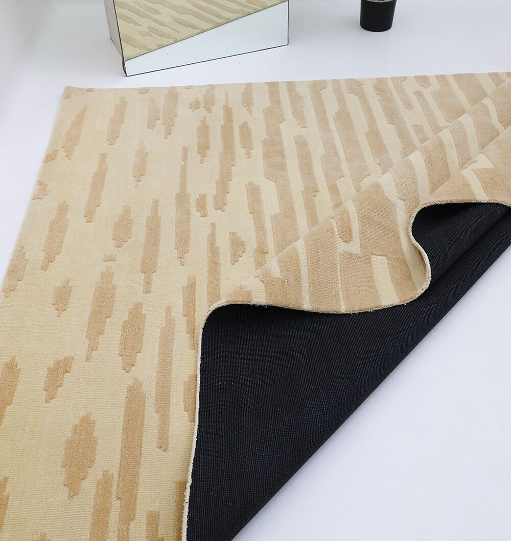 The RAMSHA HOME Dark Beige Home Decor Rug - CMN 88 (200x300 cm) is partially flipped to show its black non-slip backing. A rectangular mirror and a black cylindrical object are placed nearby on a white surface.
