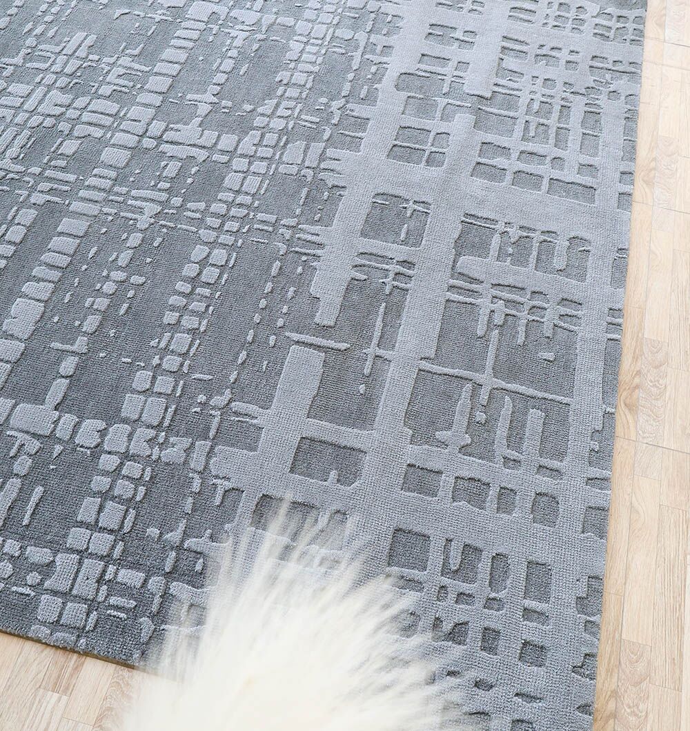The Decorative Grey Color Novara Rug - CMN 89 (200x300 cm) by RAMSHA HOME features an abstract geometric design in grey, displayed on a light wood floor and partially covered by a fluffy white item in the lower left corner.